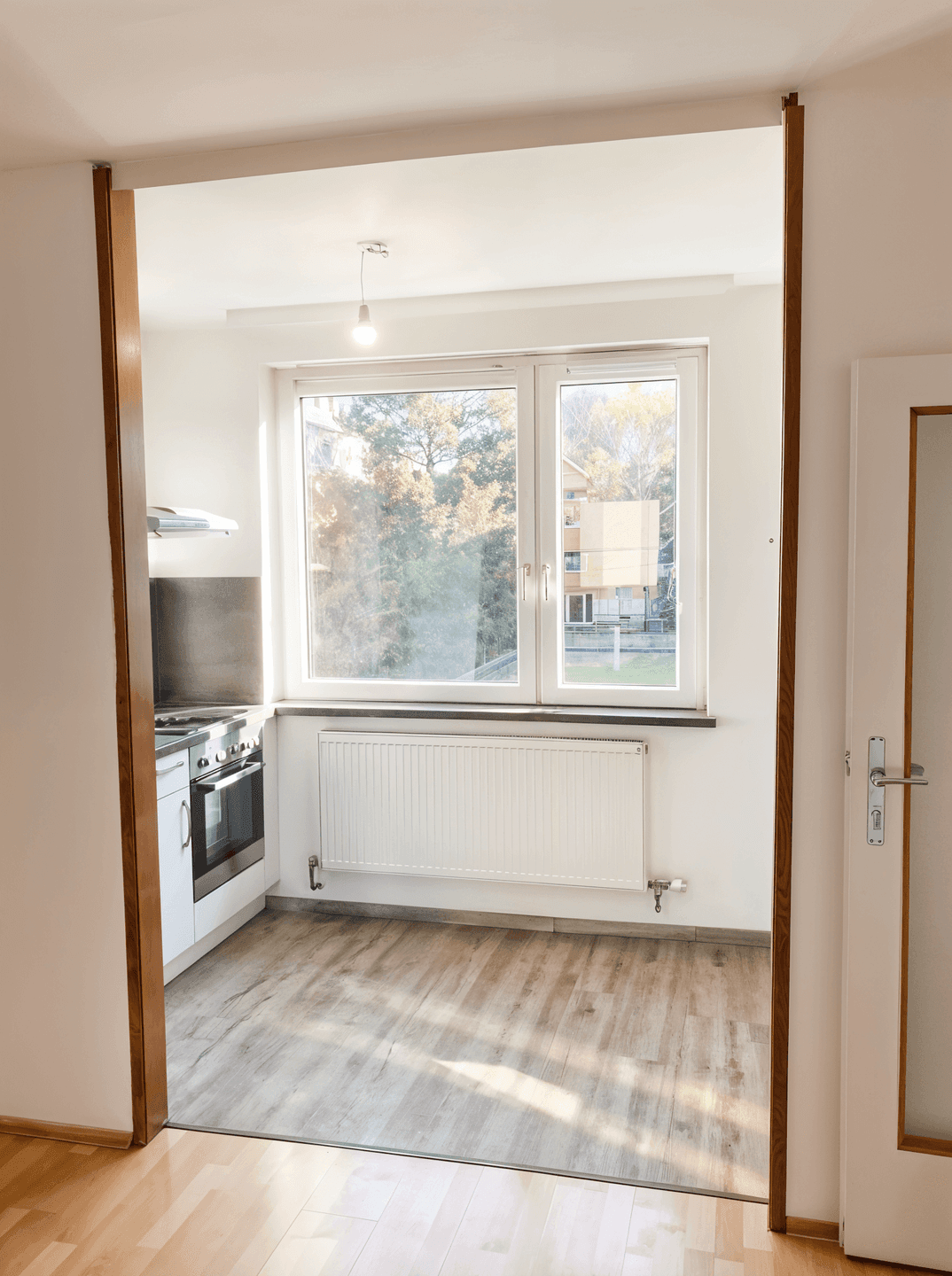 Helle Küchennische mit Fenster und Heizkörper, die durch einen Holztürrahmen vom Wohnbereich abgetrennt ist.