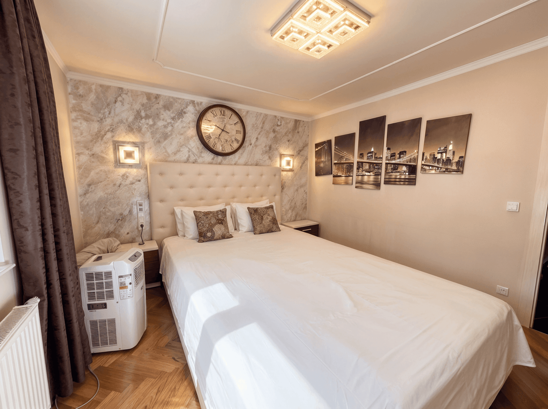 Schlafzimmer mit großem Bett, Parkettboden und mobiler Klimaanlage am Fenster.
