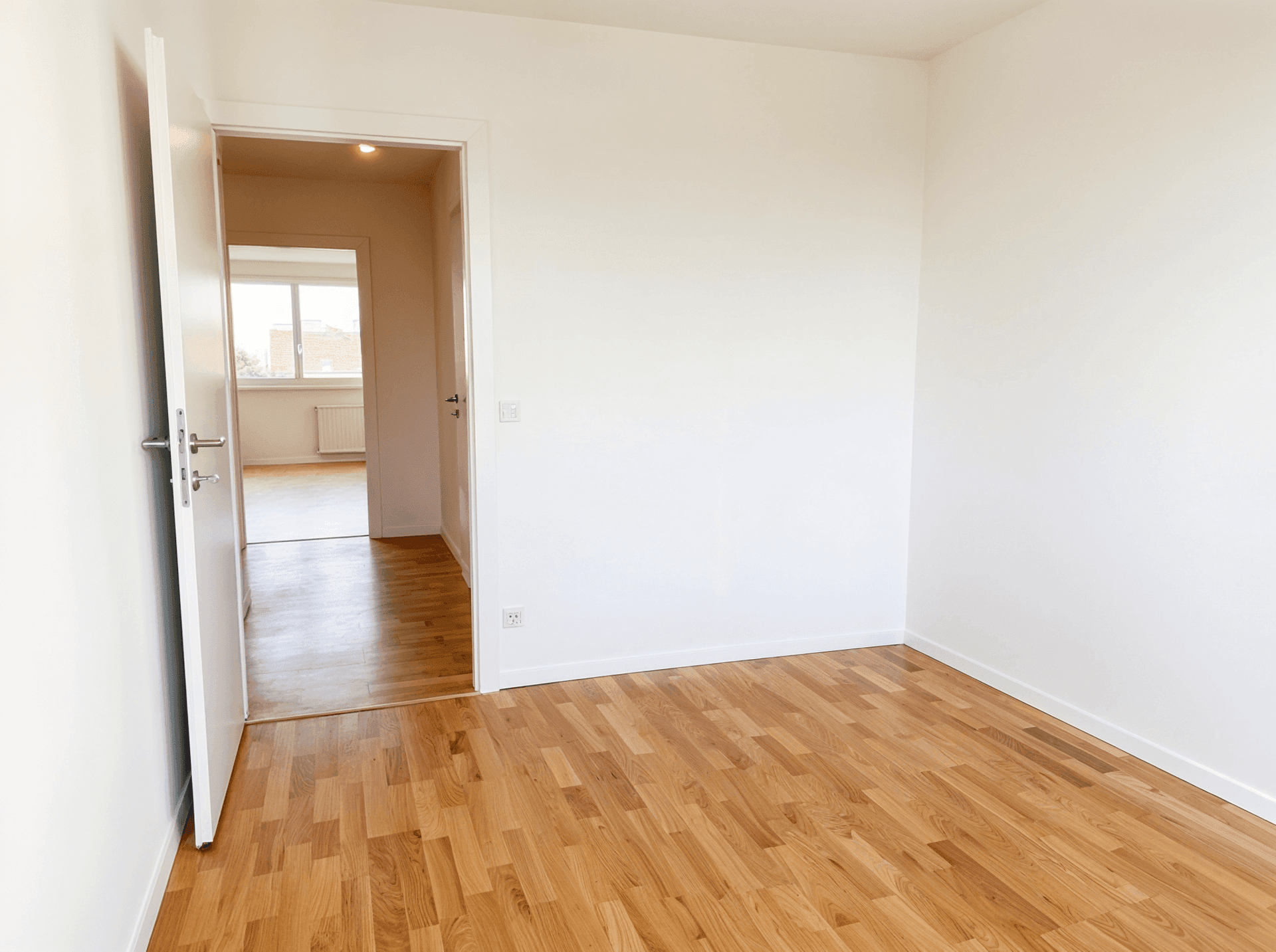 Helles Zimmer mit Parkettboden und Zugang zu einem weiteren Raum, viel Tageslicht.