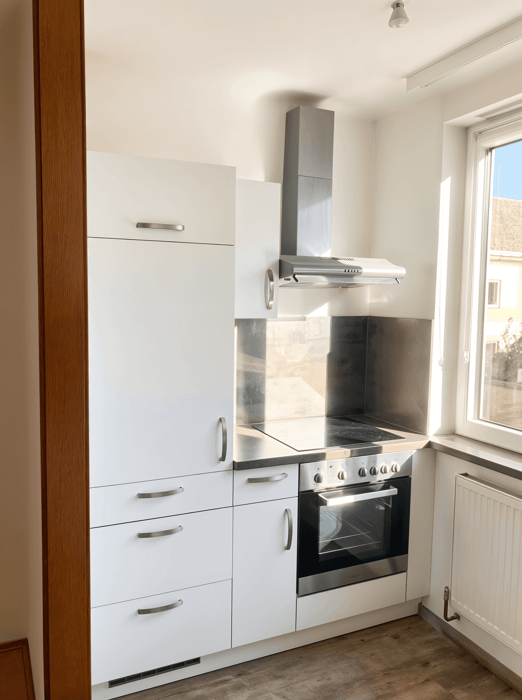 Moderne Küchenzeile mit weißen Schränken, integriertem Backofen und Dunstabzugshaube unter dem Fenster.