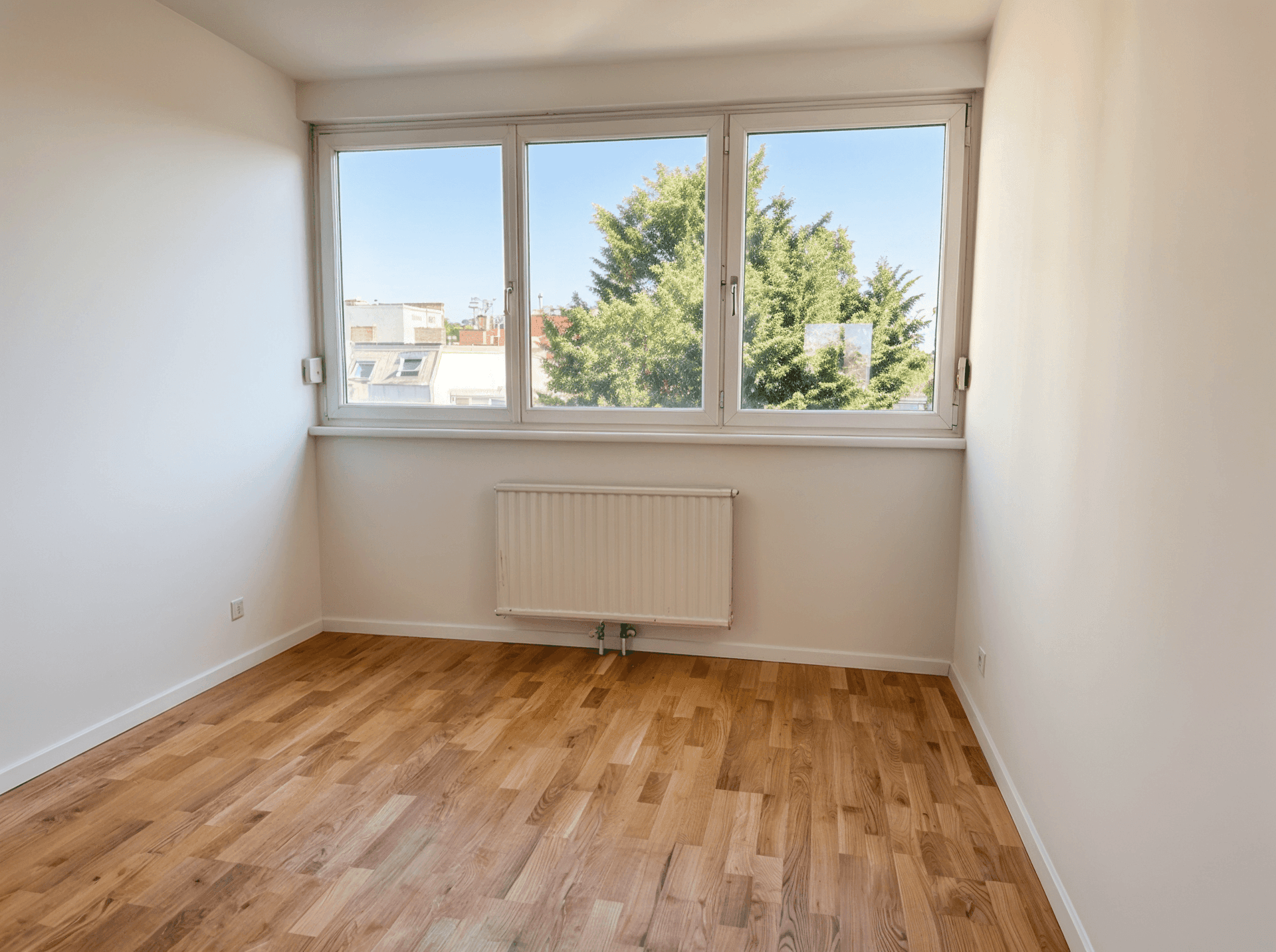 Unmöbliertes Zimmer mit großen Fenstern, Parkettboden und einem angenehmen Blick ins Grüne.
