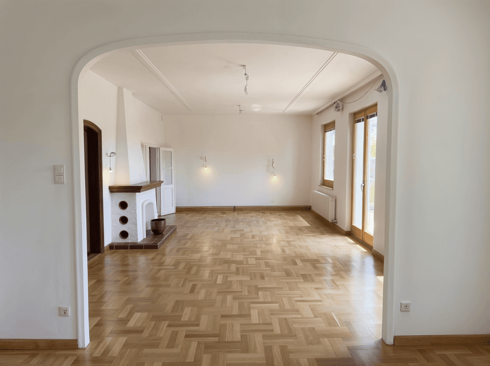 Helles Wohnzimmer mit klassischem Fischgrätparkett und einem traditionellen Kamin, verbunden durch einen Bogen.