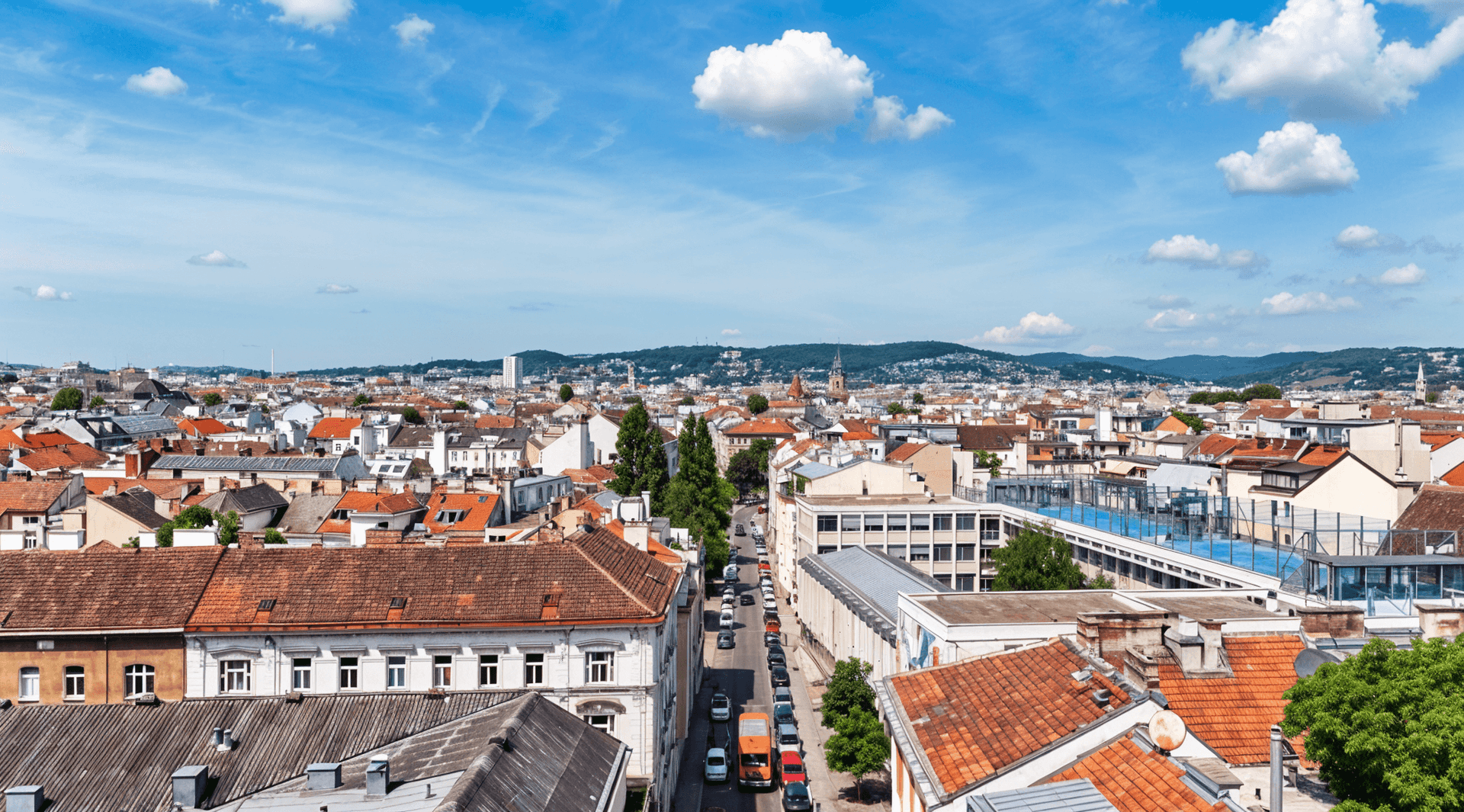 Panoramablick über eine Stadt mit zahlreichen Dächern und grünen Hügeln im Hintergrund unter blauem Himmel.