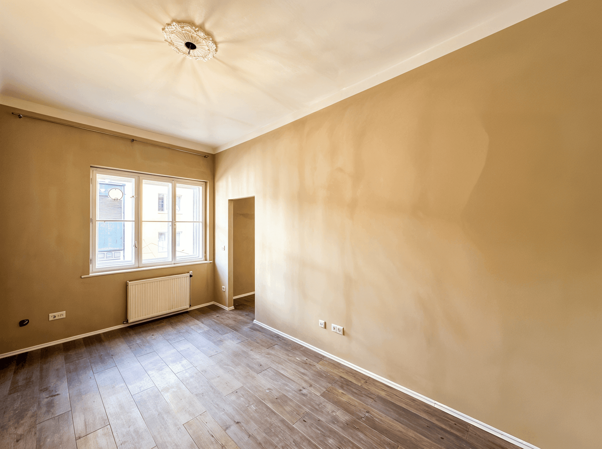 Geräumiges, unmöbliertes Zimmer mit Holzboden, großem Fenster und einem kleinen Wanddurchbruch.