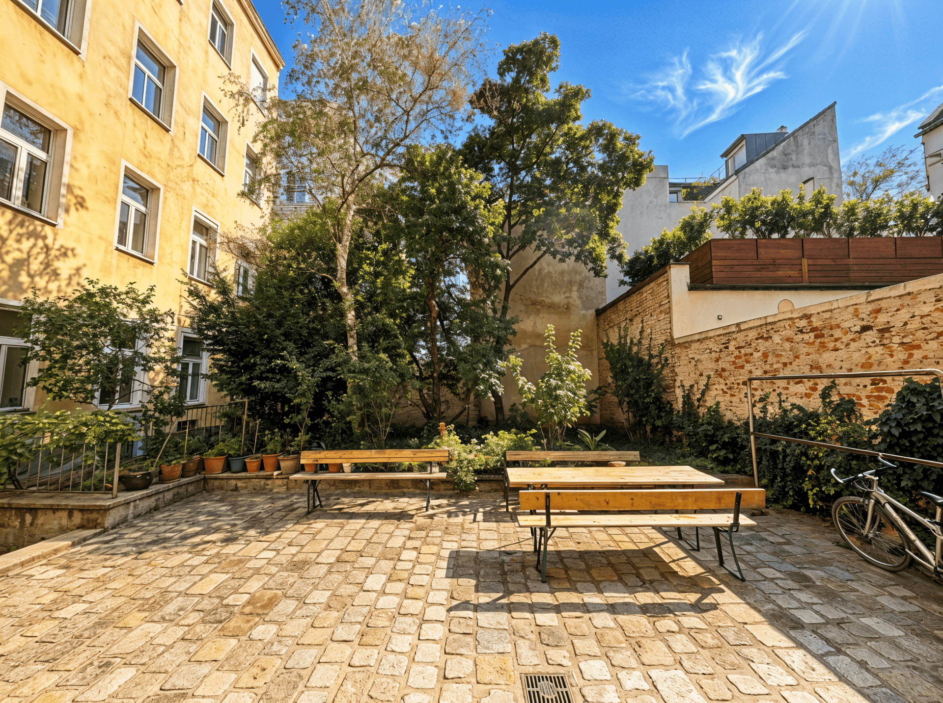 Sonniger Innenhof mit Sitzgelegenheiten, Bäumen und gepflastertem Boden, umgeben von Gebäuden.