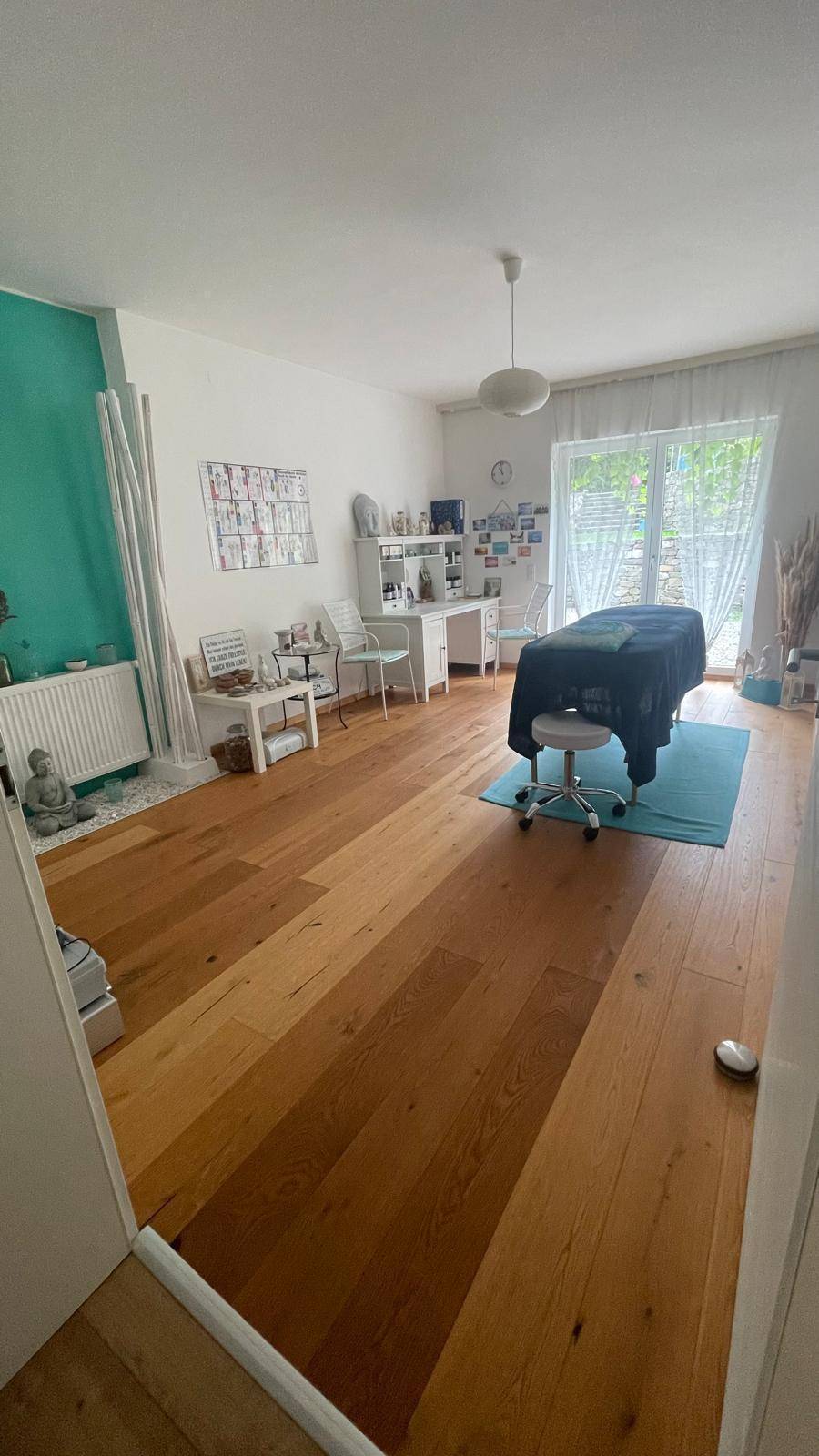 Vielseitiger Raum mit Holzboden, türkiser Akzentwand und großem Fenster für natürliches Licht.