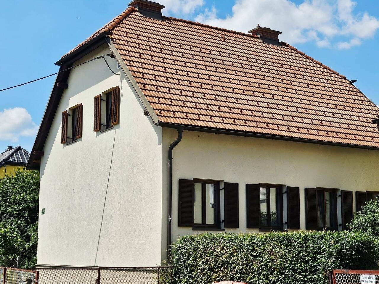 Ansicht der Hausfassade mit Ziegeldach, braunen Fensterläden und einer grünen Hecke.