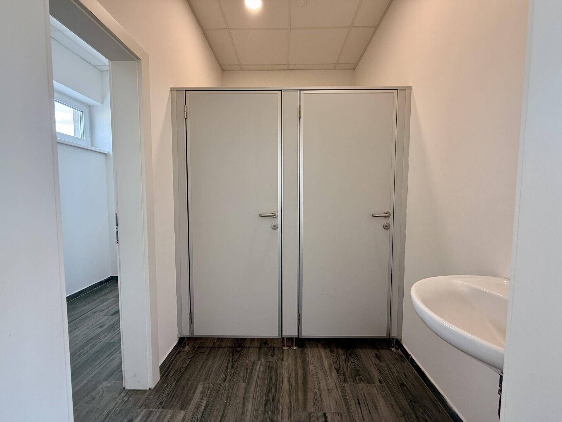 Moderner WC-Bereich mit zwei Kabinen, einem Waschbecken und dunklem Fliesenboden.