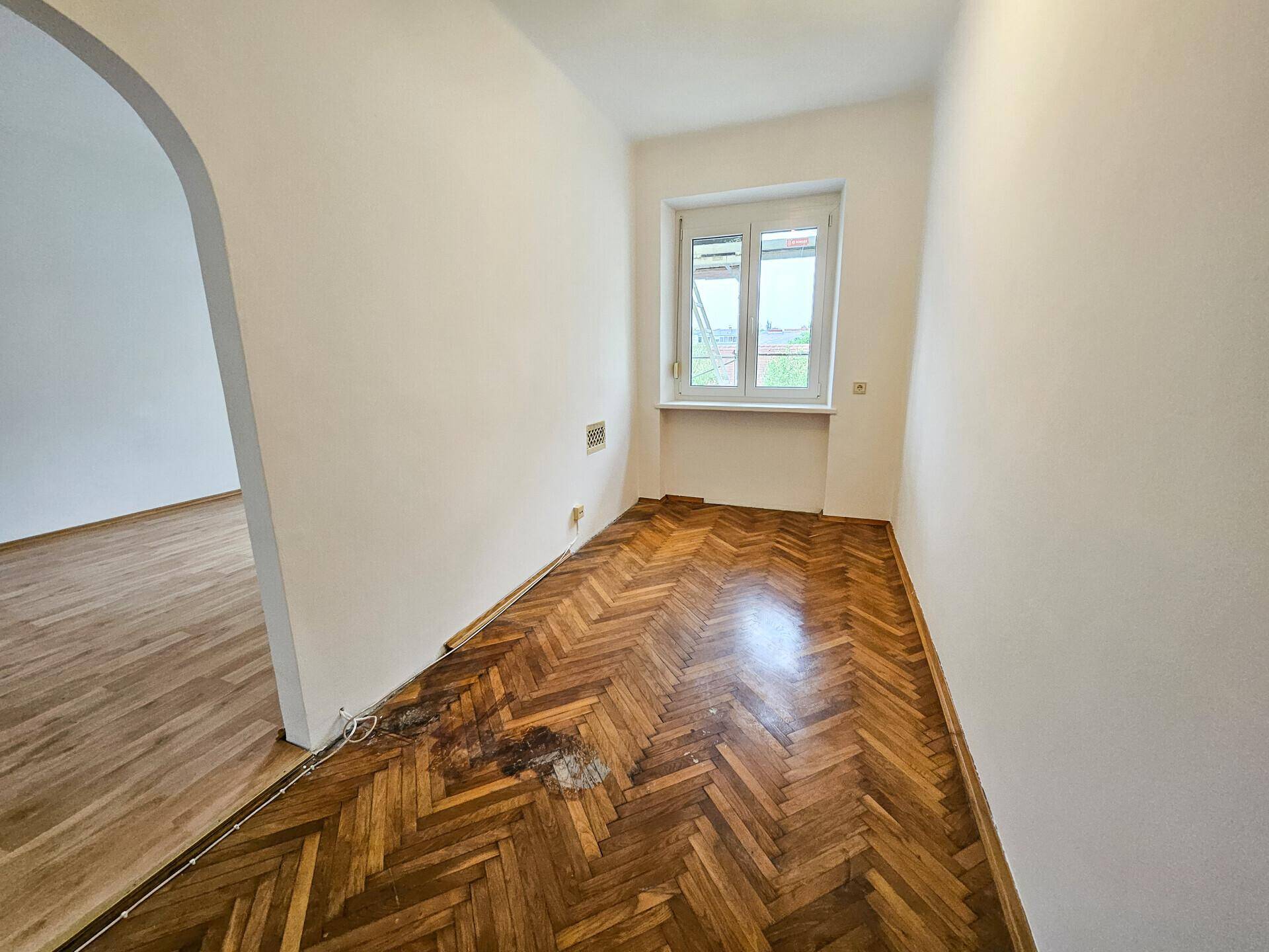 Kleineres Zimmer mit Fischgrätparkett, Fenster und einem offenen Durchgang zum Wohnbereich.