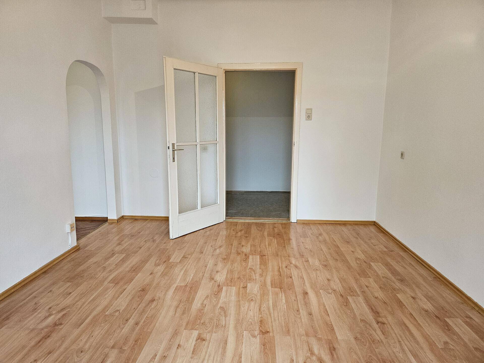 Heller Raum mit Holzboden, einer Tür und einem offenen Durchgang zu einem weiteren Bereich.