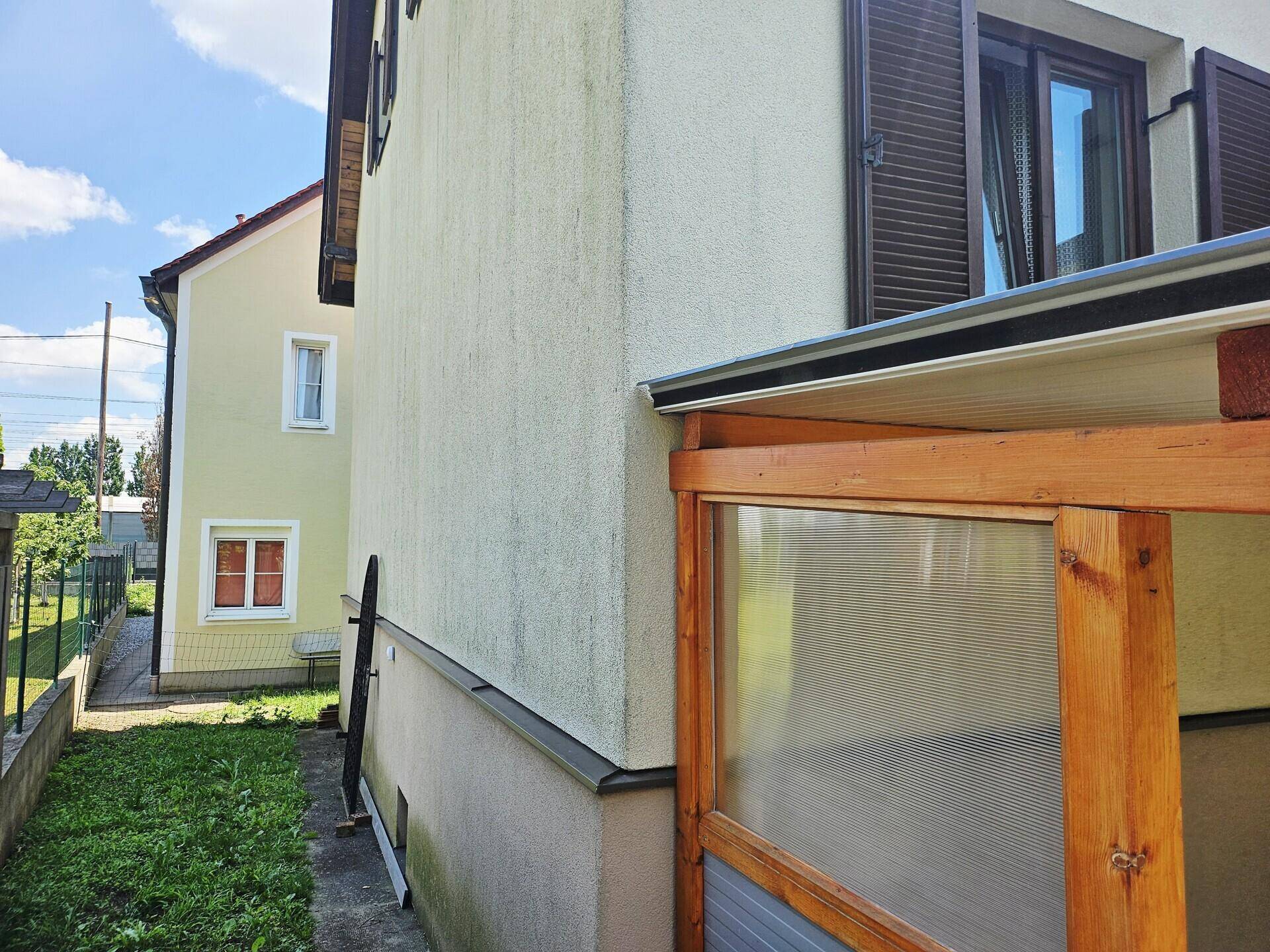 Nahaufnahme der Hauswand mit angrenzendem Holzunterstand und Blick auf den Gartenbereich.