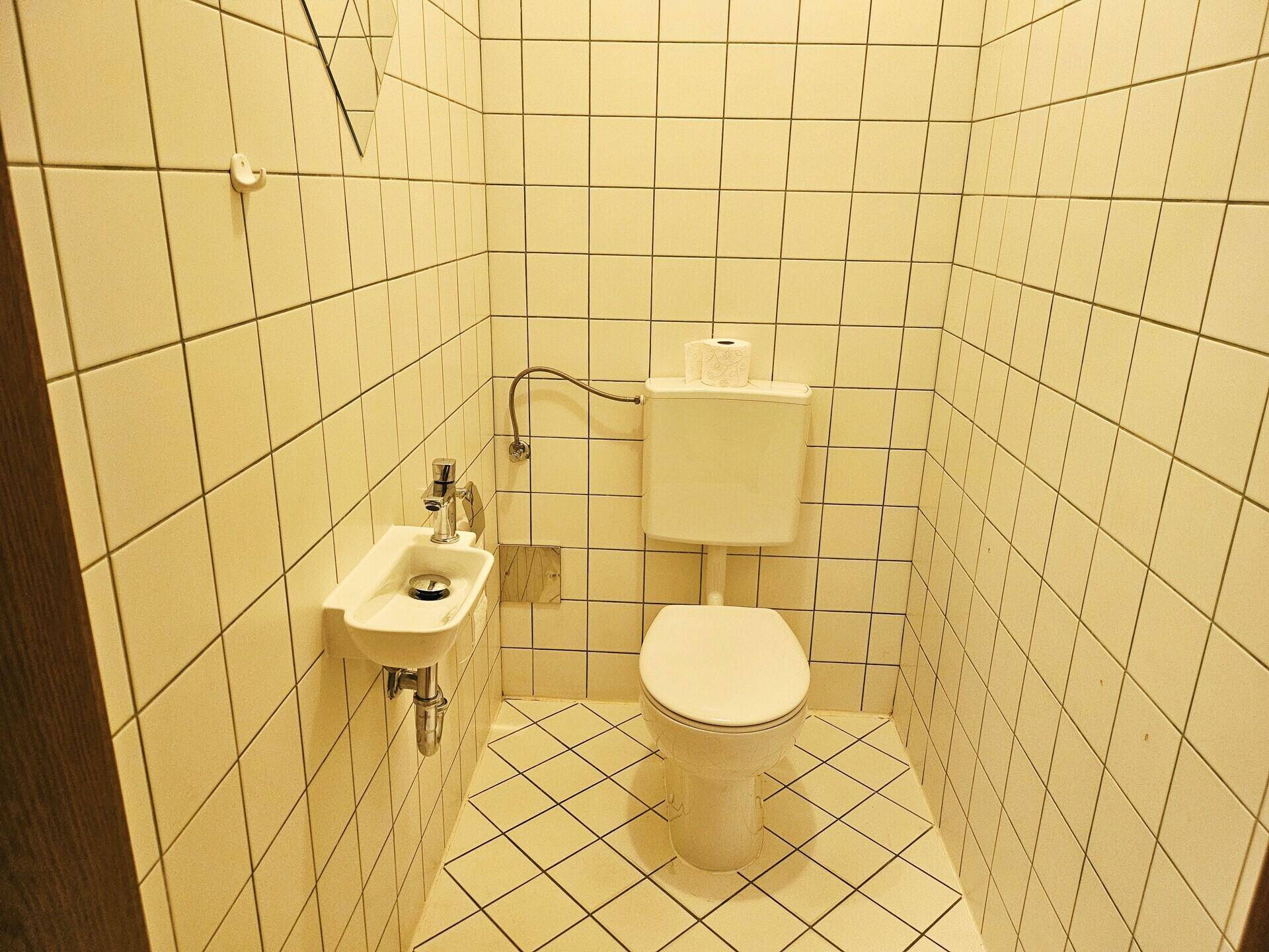 Kleines Gäste-WC mit weißen Fliesen, einer Toilette und einem kompakten Handwaschbecken.