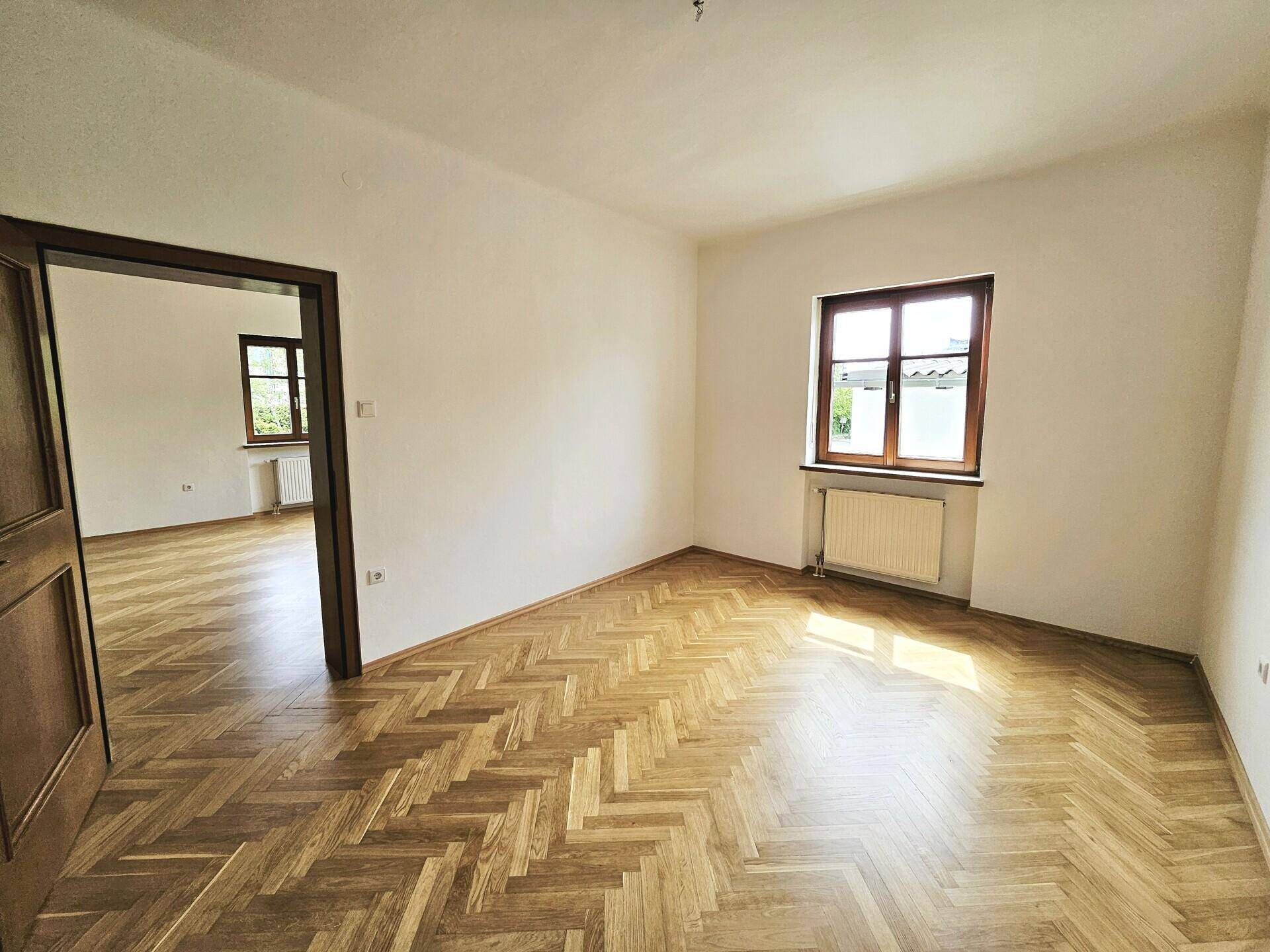 Helles Wohnzimmer mit klassischem Fischgrätparkett und einem Durchgang zu einem weiteren Raum.