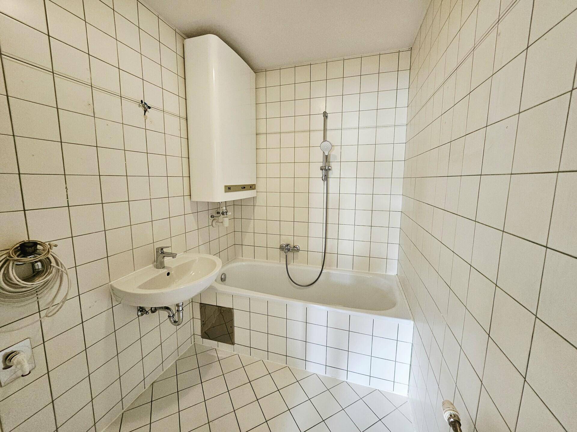 Gefliestes Badezimmer mit Badewanne, Duscharmatur und einem einfachen Waschbecken in weißer Keramik.