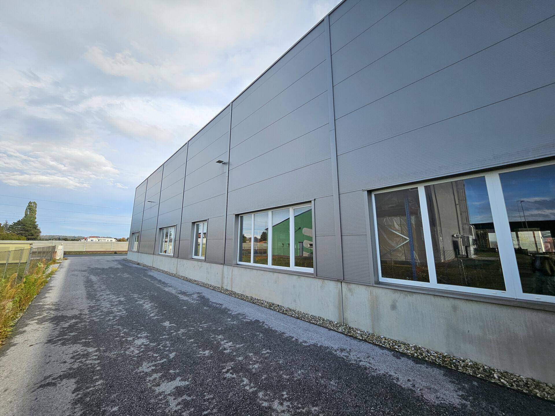 Lange Seitenansicht des Industriegebäudes mit grauer Fassade, Reihen von Fenstern und einer gepflasterten Zufahrt.