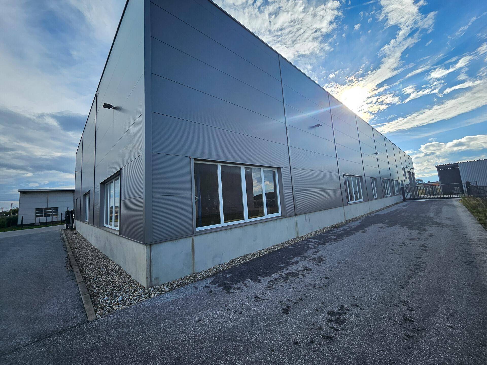 Seitenansicht des modernen Industriegebäudes mit grauer Fassade und mehreren Fenstern unter sonnigem Himmel.