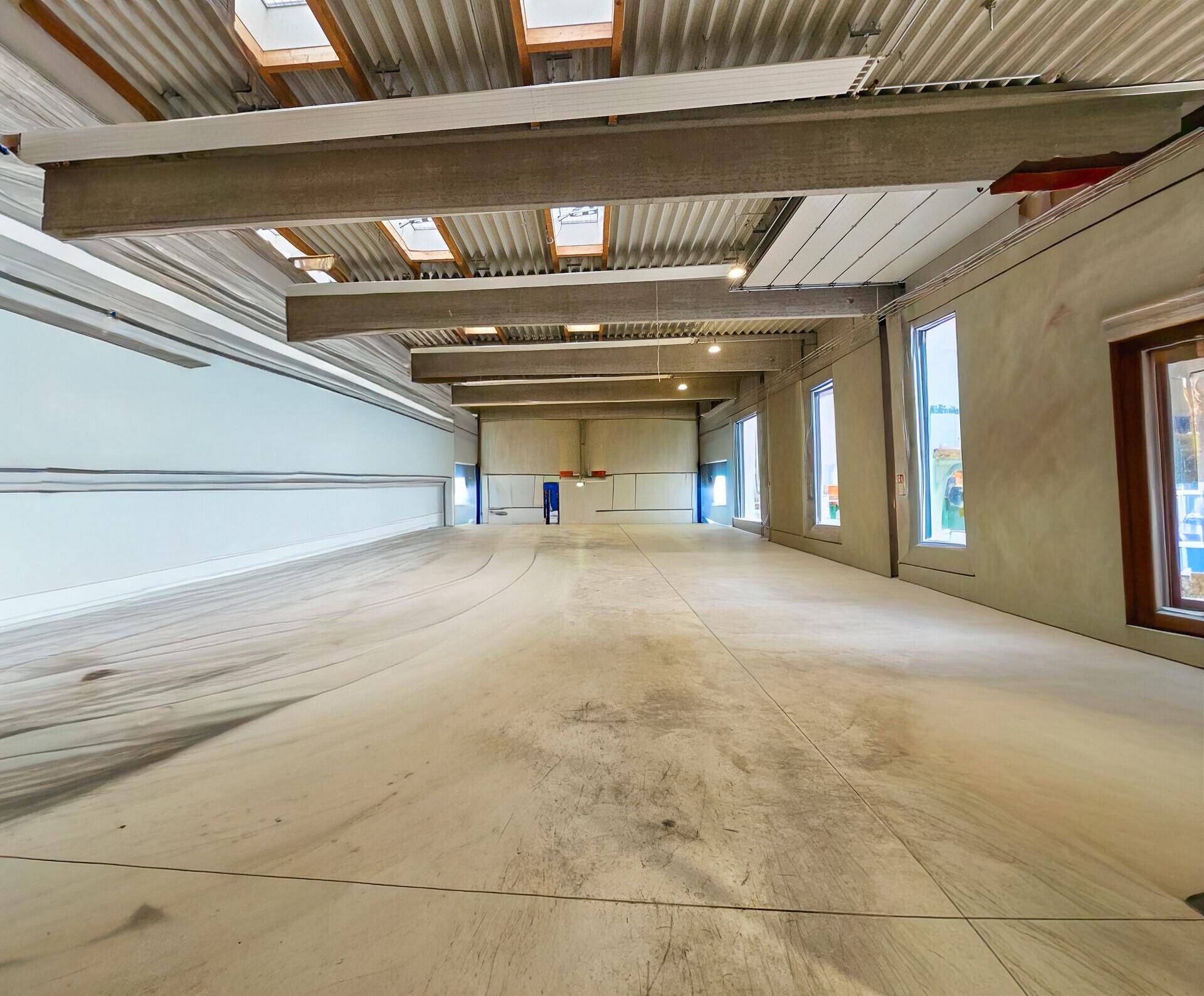 Geräumige Industriehalle mit hohen Decken, sichtbaren Trägern, Betonboden und Oberlichtern für viel Tageslicht.