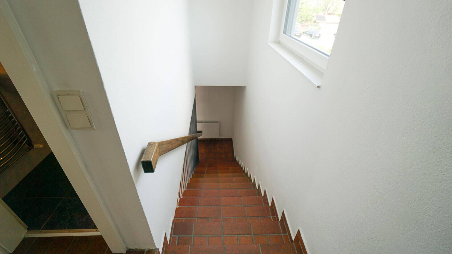 Treppenhaus mit Terrakotta-Fliesenstufen und weiß gestrichenen Wänden, gut beleuchtet durch ein Fenster.