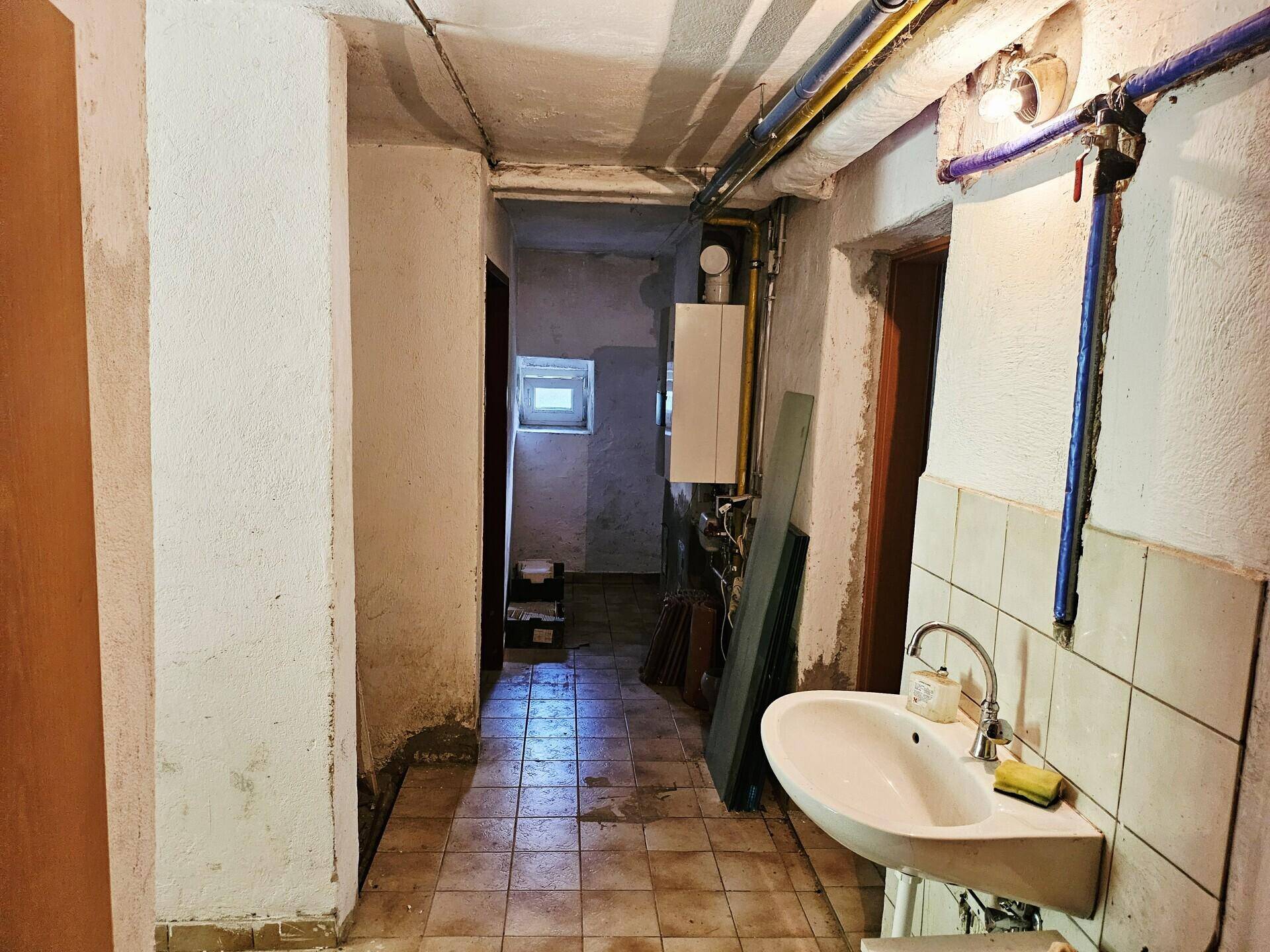 Kellerflur mit Waschbecken, sichtbaren Rohren und einfacher Ausstattung.