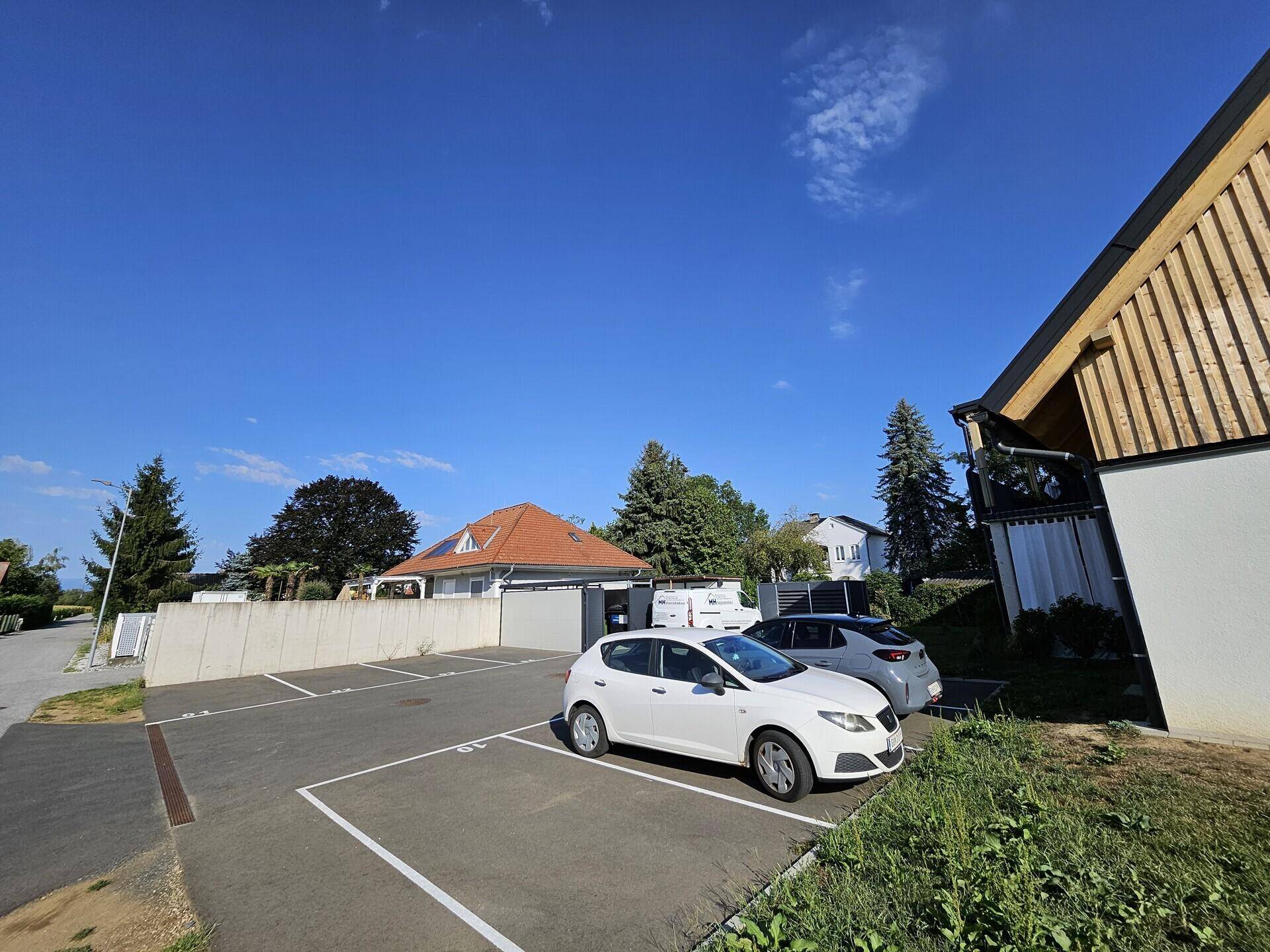 Außenansicht mit Parkplätzen und einem Teil der modernen Wohngebäude unter strahlend blauem Himmel.