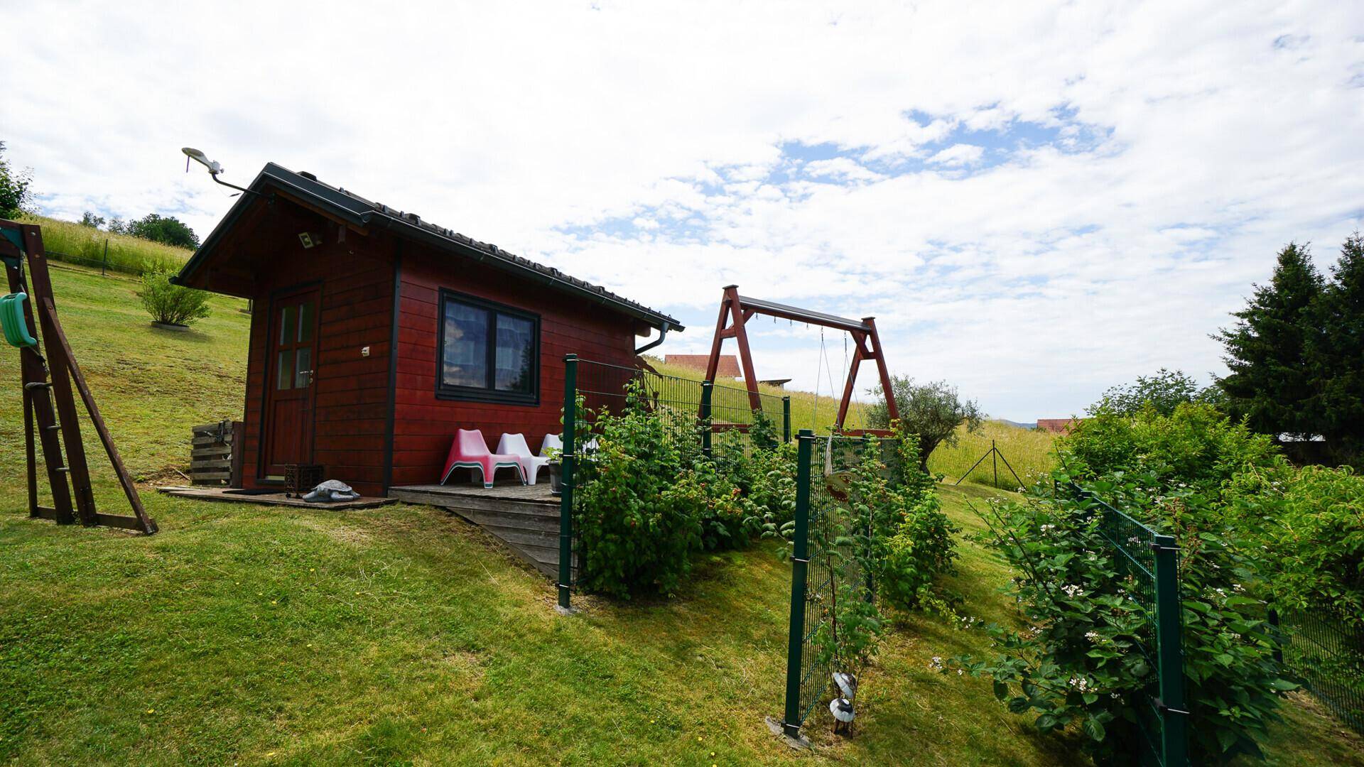 Kleines Gartenhaus aus Holz mit Terrasse und Schaukel im weitläufigen, grünen Gartenbereich.