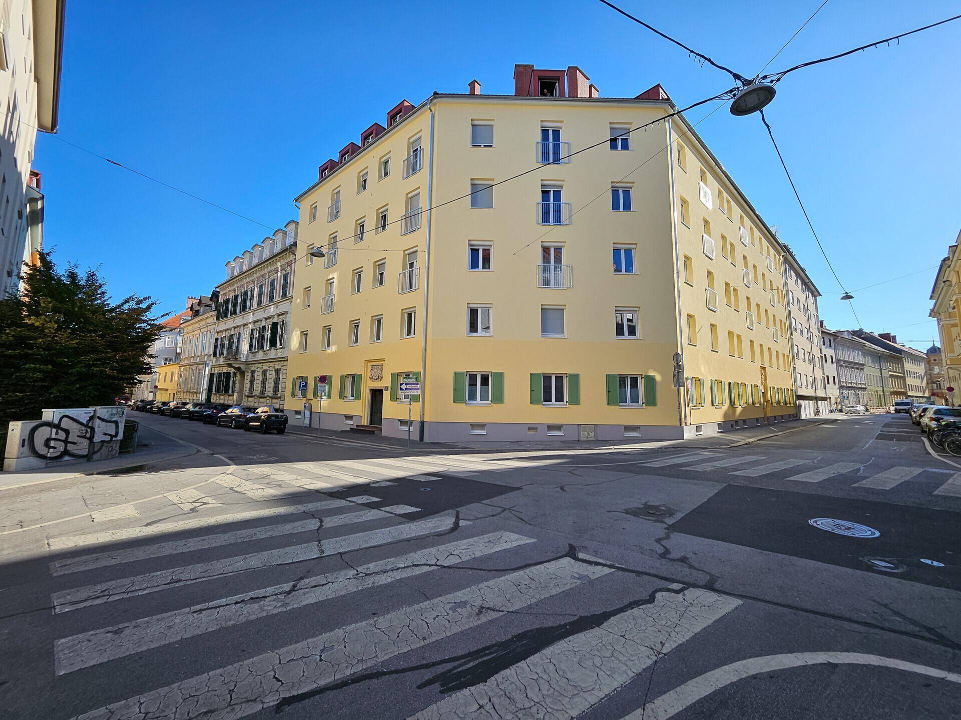 Eckgebäude mit hellgelber Fassade, grünen Fensterläden und kleinen Balkonen an einem sonnigen Tag.