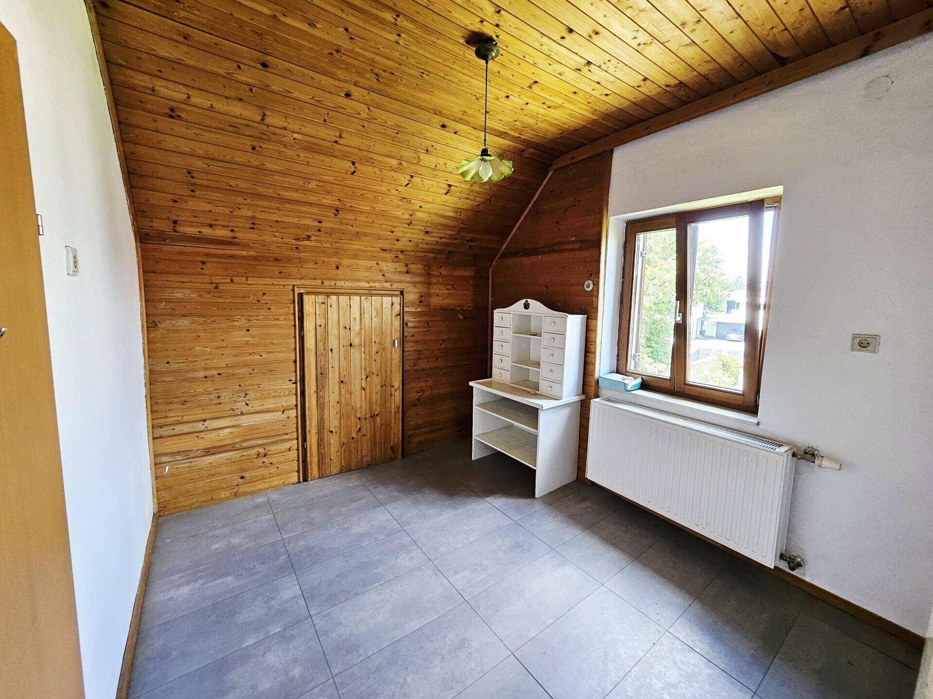 Dachzimmer mit Holzvertäfelung, Fliesenboden und einem Fenster mit Ausblick.