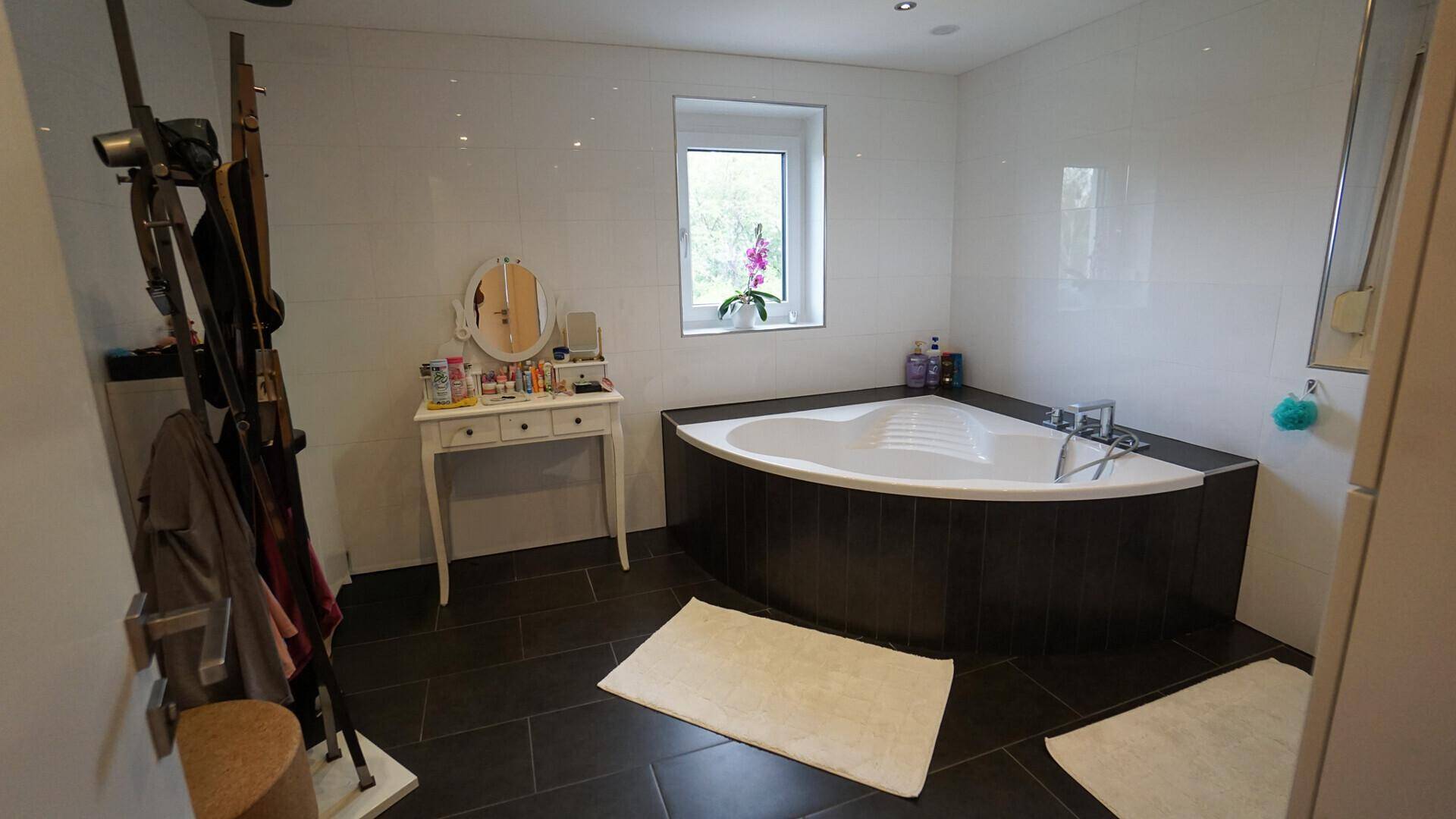 Geräumiges Badezimmer mit moderner Eckbadewanne, dunklen Fliesen und einem Fenster für Tageslicht.