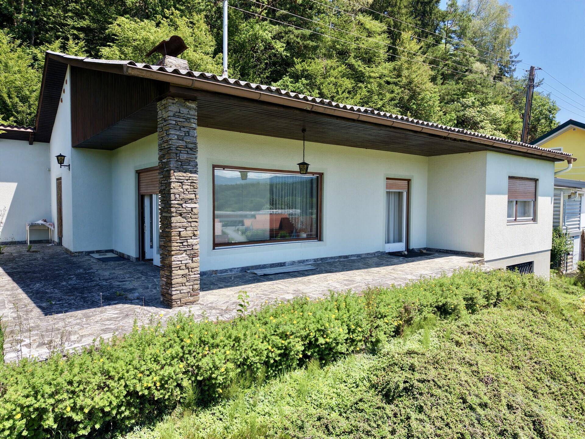 Fassade des Hauses mit Steinpfeiler und großer Fensterfront, umgeben von Sträuchern.