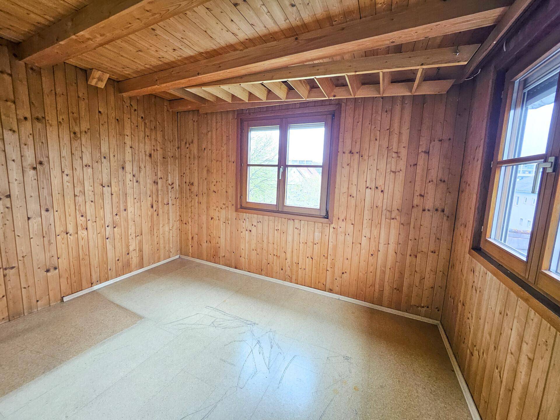 Zimmer mit Holzwänden, Holzdecke und zwei Fenstern für viel Tageslicht.
