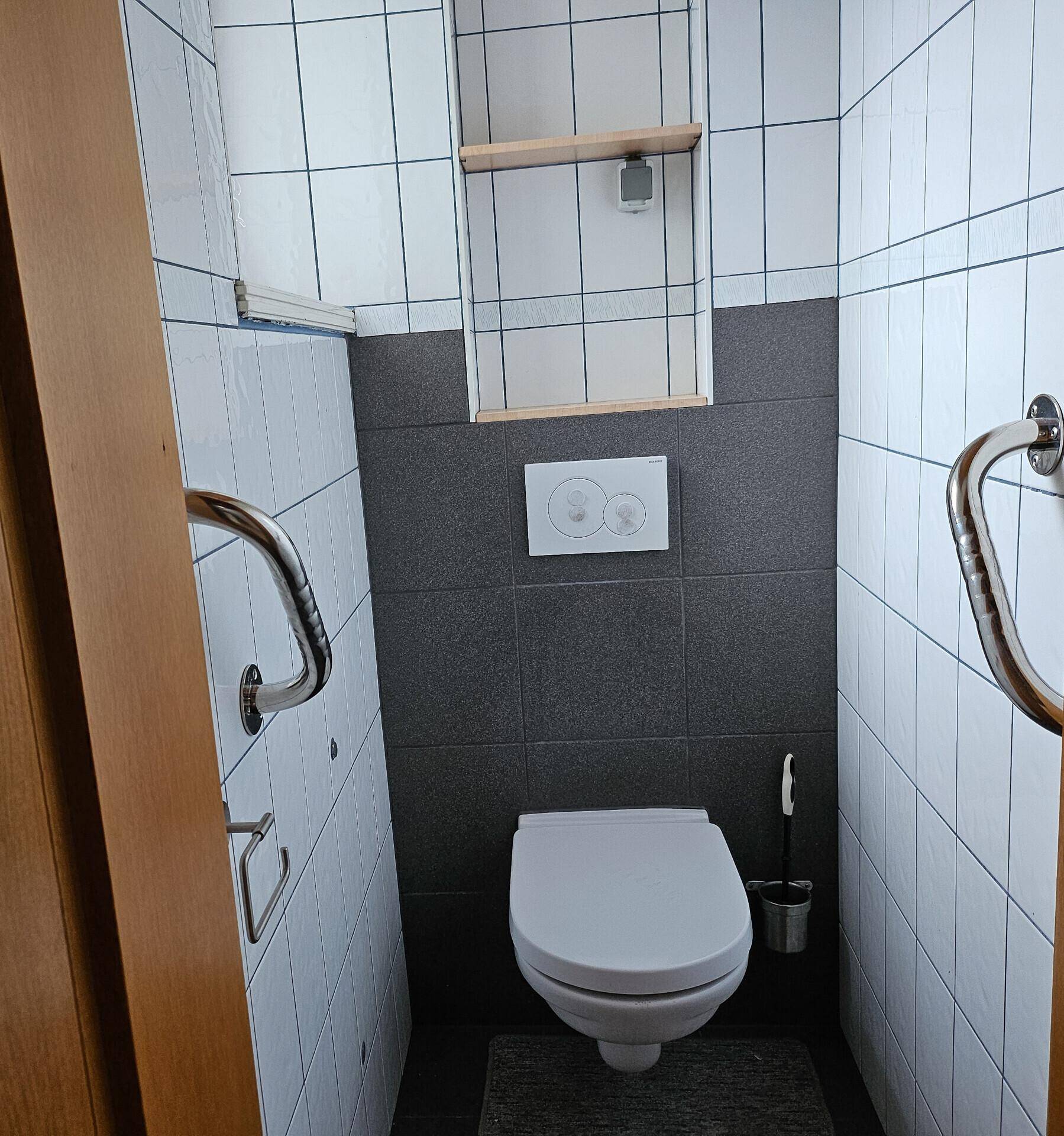 Modernisiertes Gäste-WC mit dunklen und weißen Fliesen, Wand-WC und Haltegriffen.