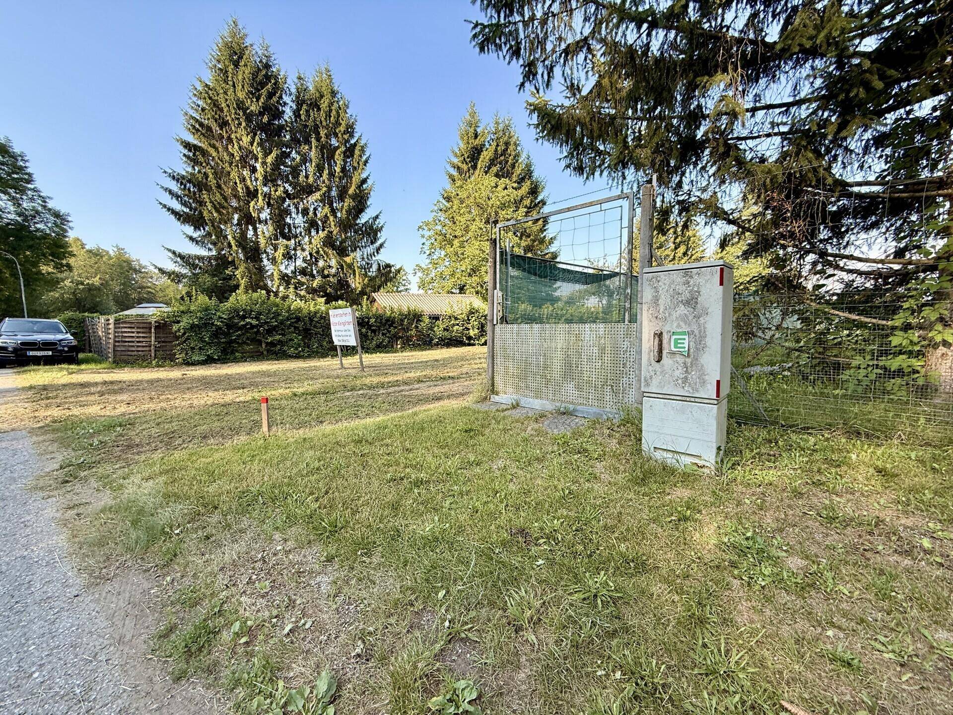 Eingangsbereich zu einem Grundstück mit Tor, Elektroverteilerkasten und umgebender Natur.