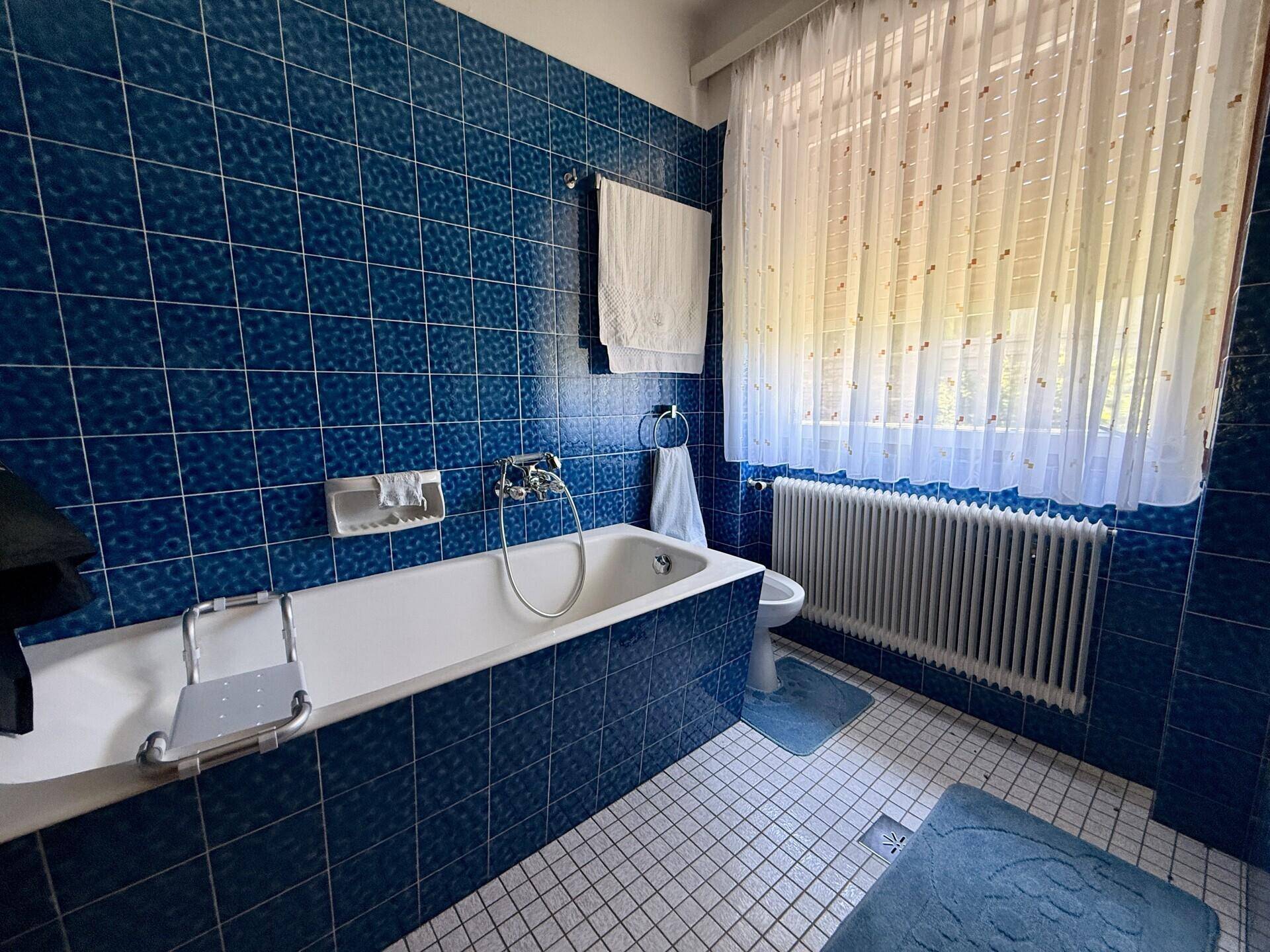 Badezimmer mit blauen Fliesen, Badewanne und Fenster, ausgestattet mit Haltegriffen.