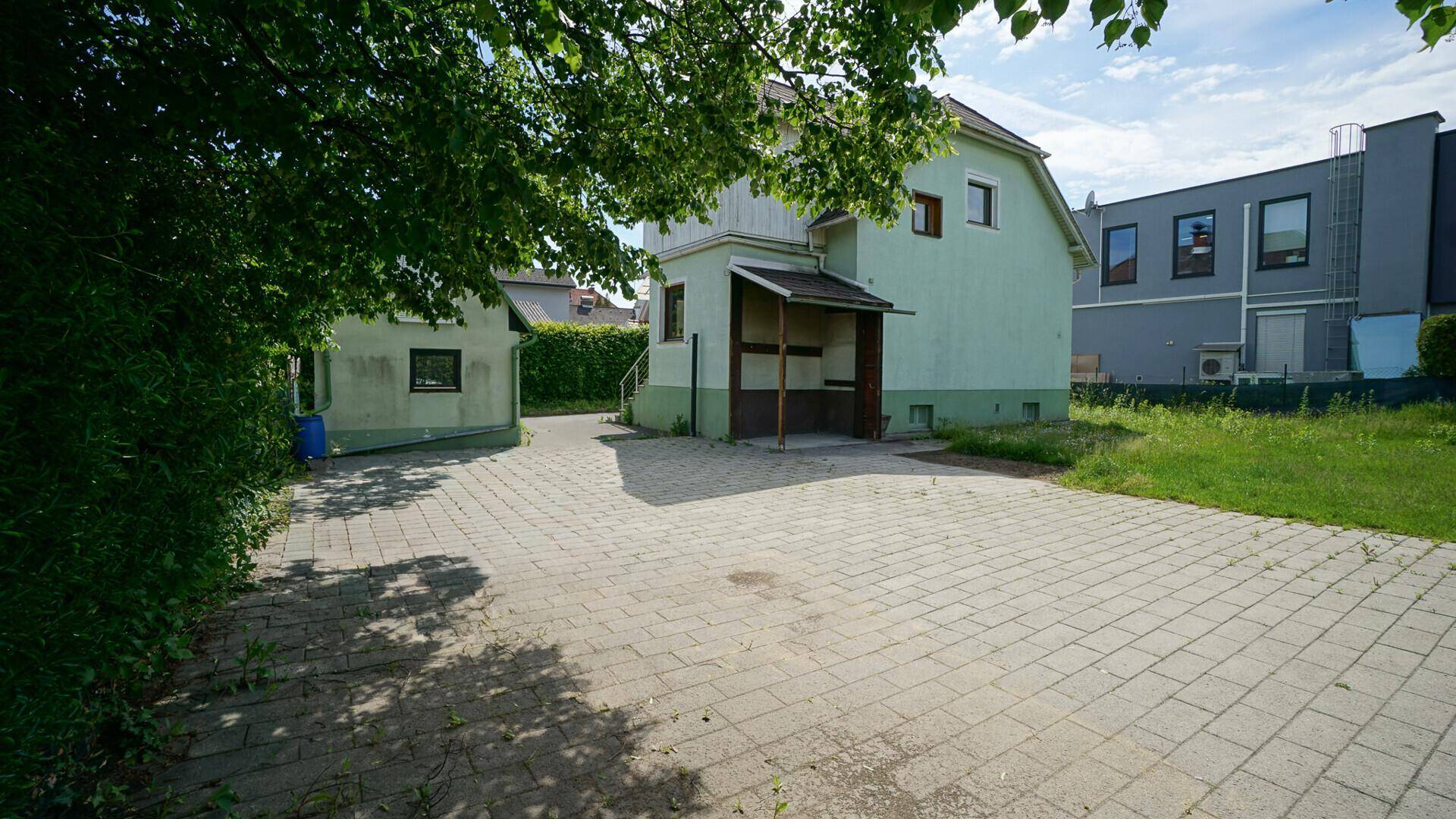 Rückseite eines hellgrünen Hauses mit gepflastertem Hof und viel Grün unter sonnigem Himmel.