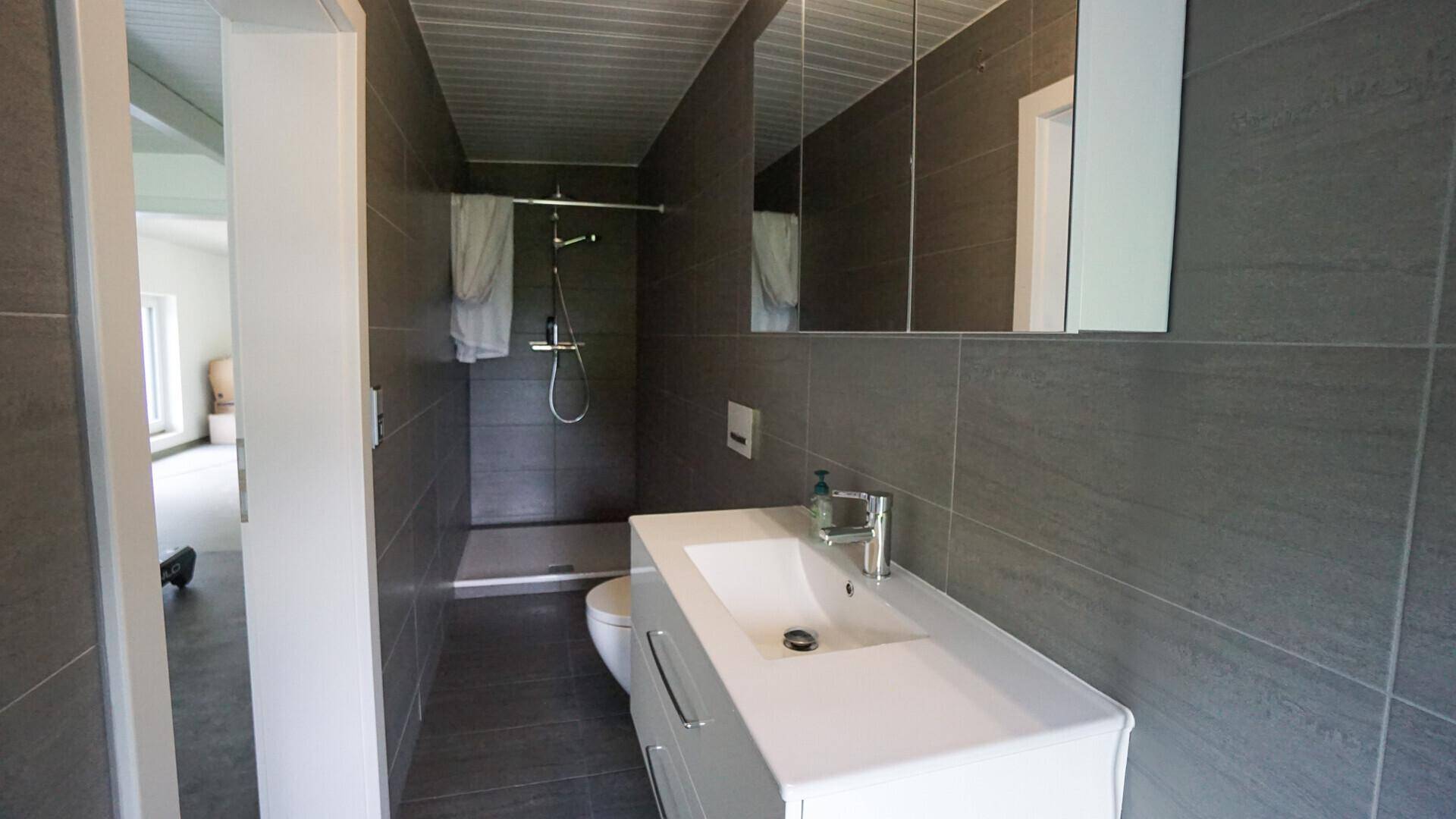Modernes Badezimmer mit grauen Großformatfliesen, Waschtisch und ebenerdiger Dusche.