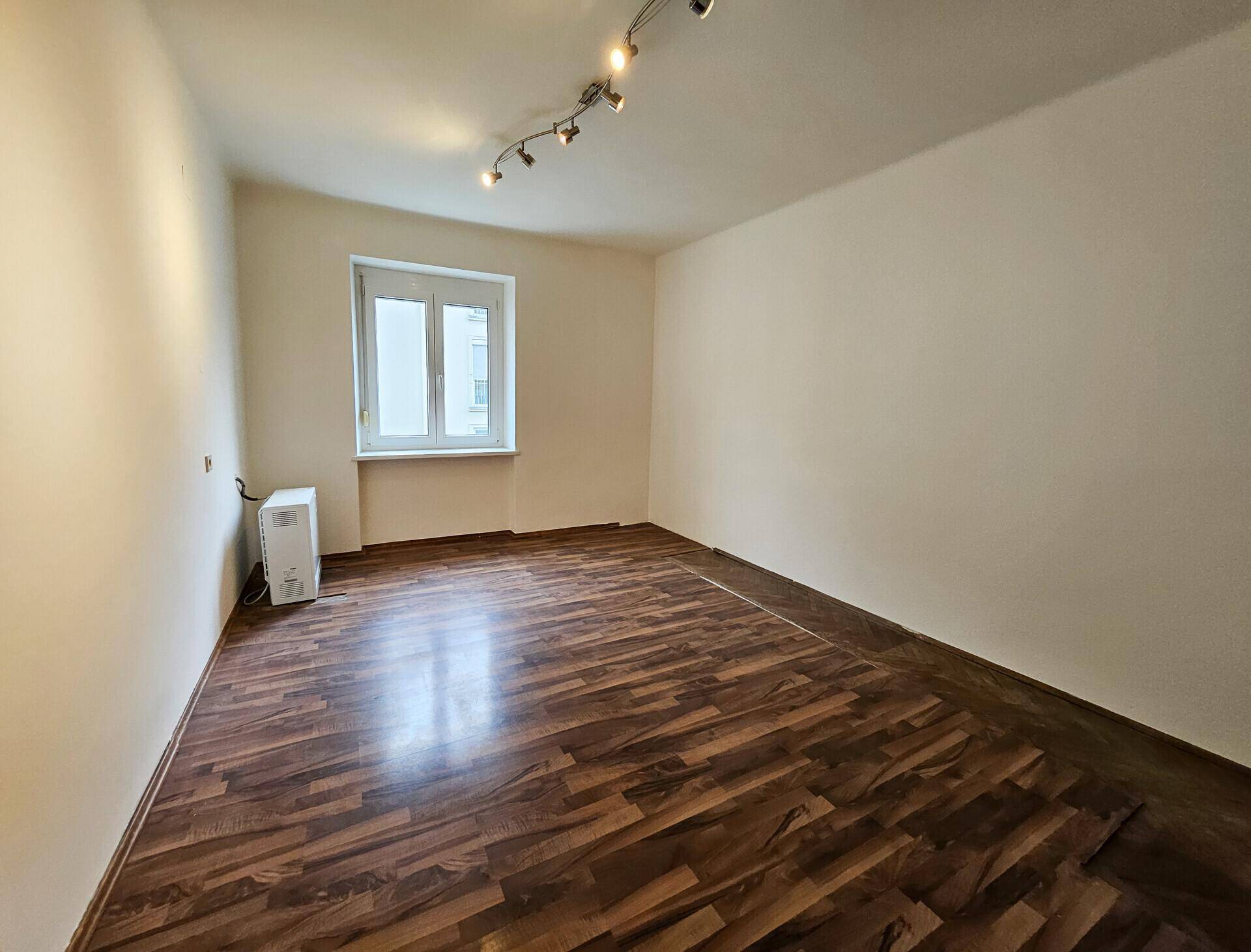 Leeres Zimmer mit Laminatboden, weiß gestrichenen Wänden und einem Fenster für Tageslicht.