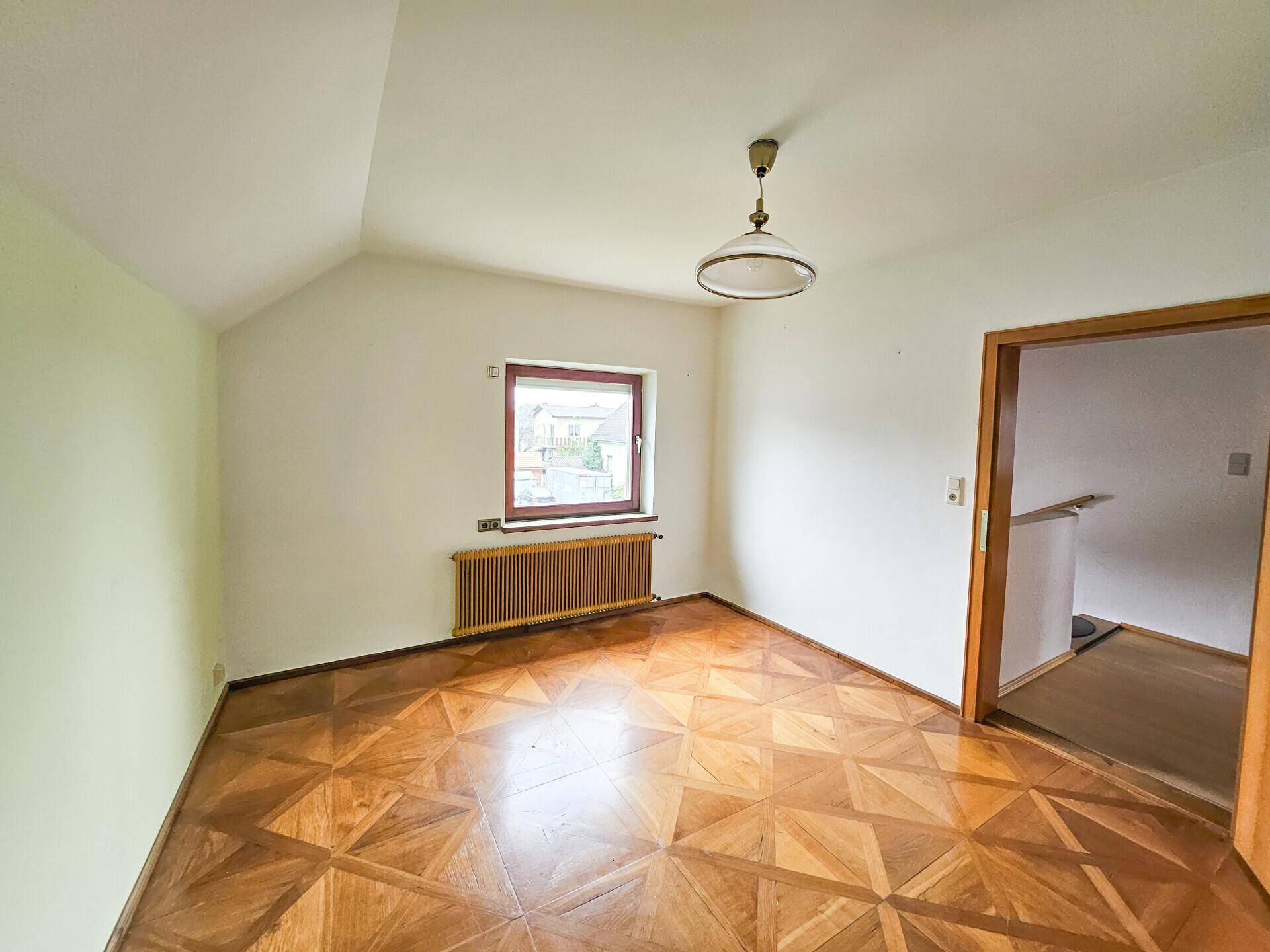 Helles Zimmer mit Parkettboden, Fenster und Zugang zu einem weiteren Raum.