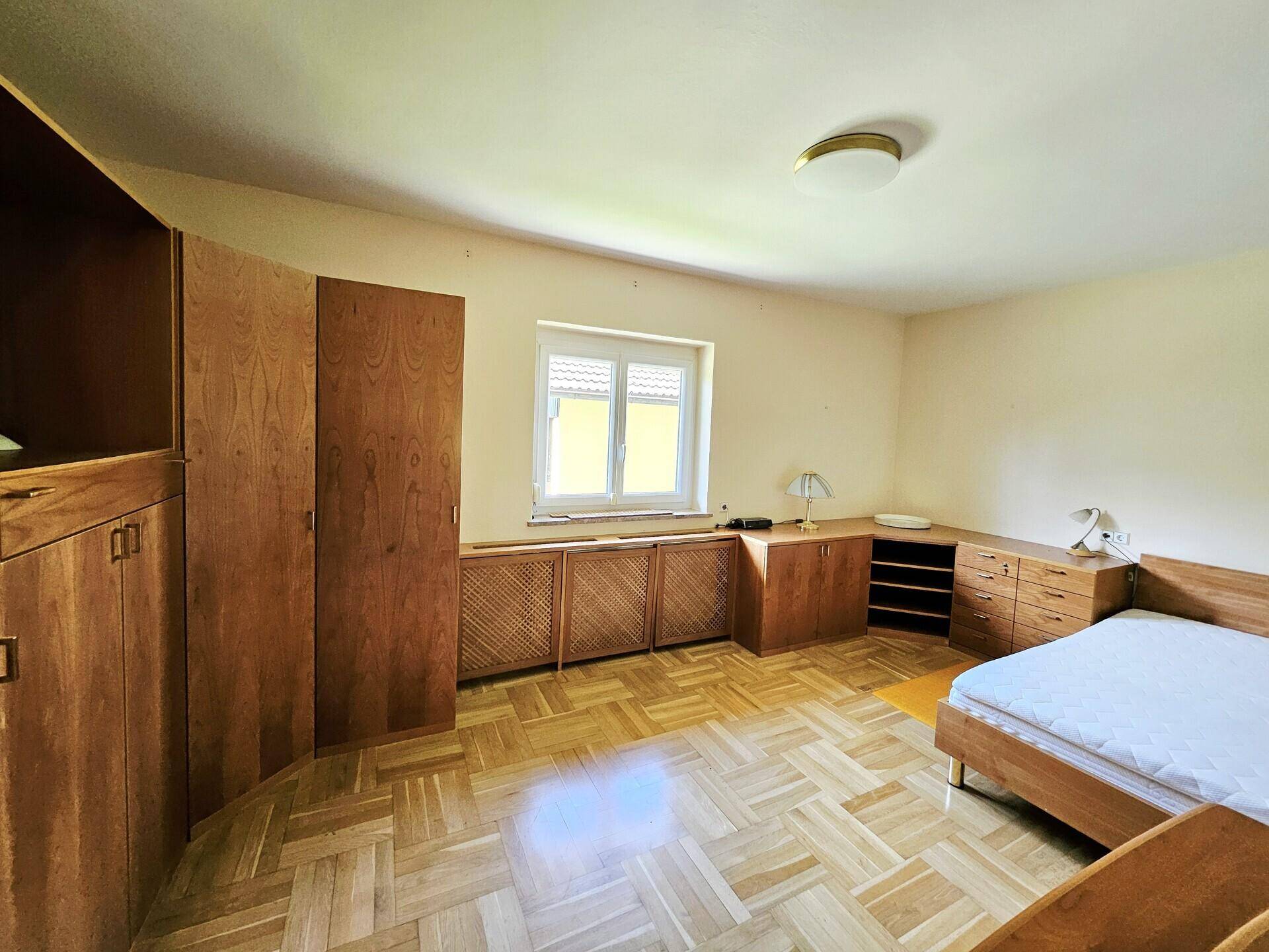 Schlafzimmer mit Parkettboden, Holzmöbeln und Fenster mit Blick ins Grüne.