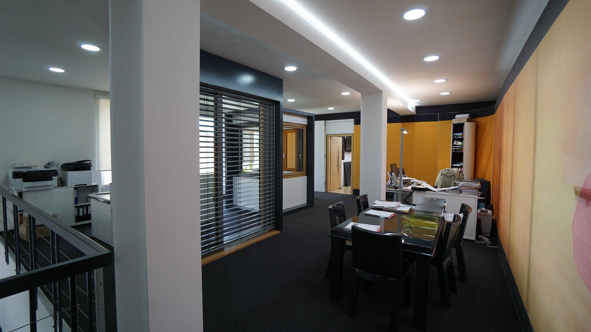 Geräumiges Büro mit Teppichboden, Besprechungstisch und Blick auf einen weiteren Arbeitsbereich.