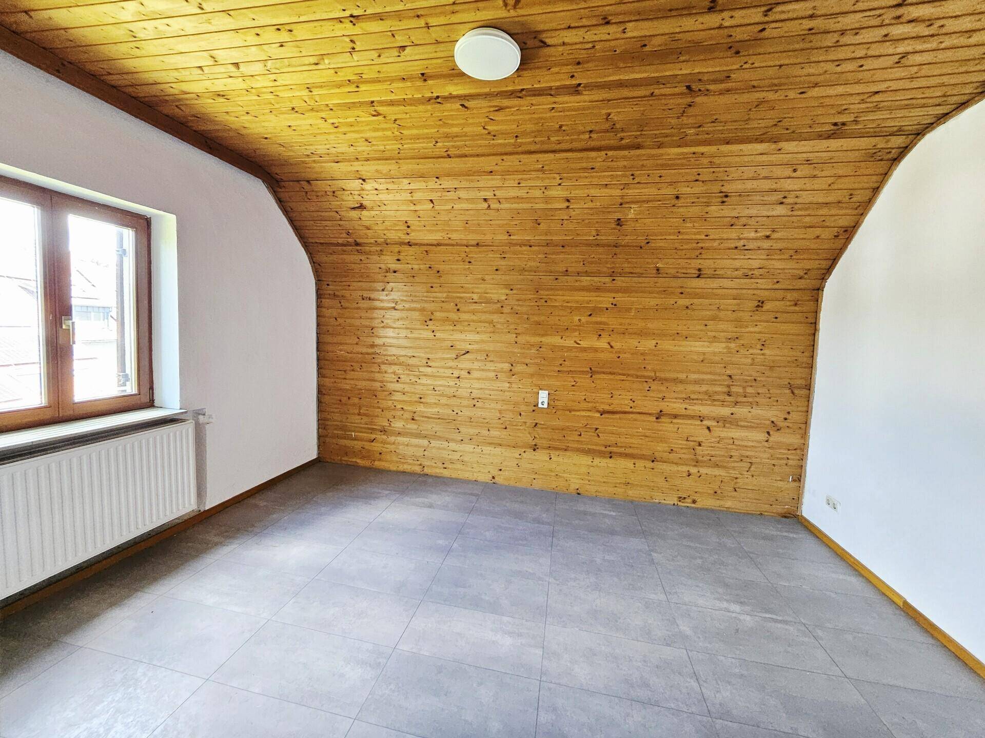 Helles Dachzimmer mit Holzdecke, Fliesenboden und einem großen Fenster.