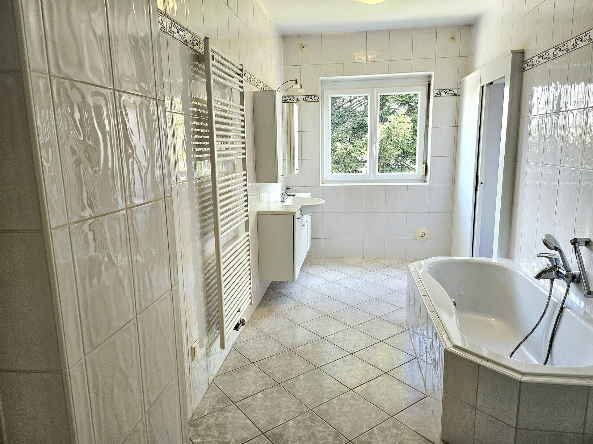 Helles Badezimmer mit Fenster, Badewanne, Duschkabine und weißen Fliesen.