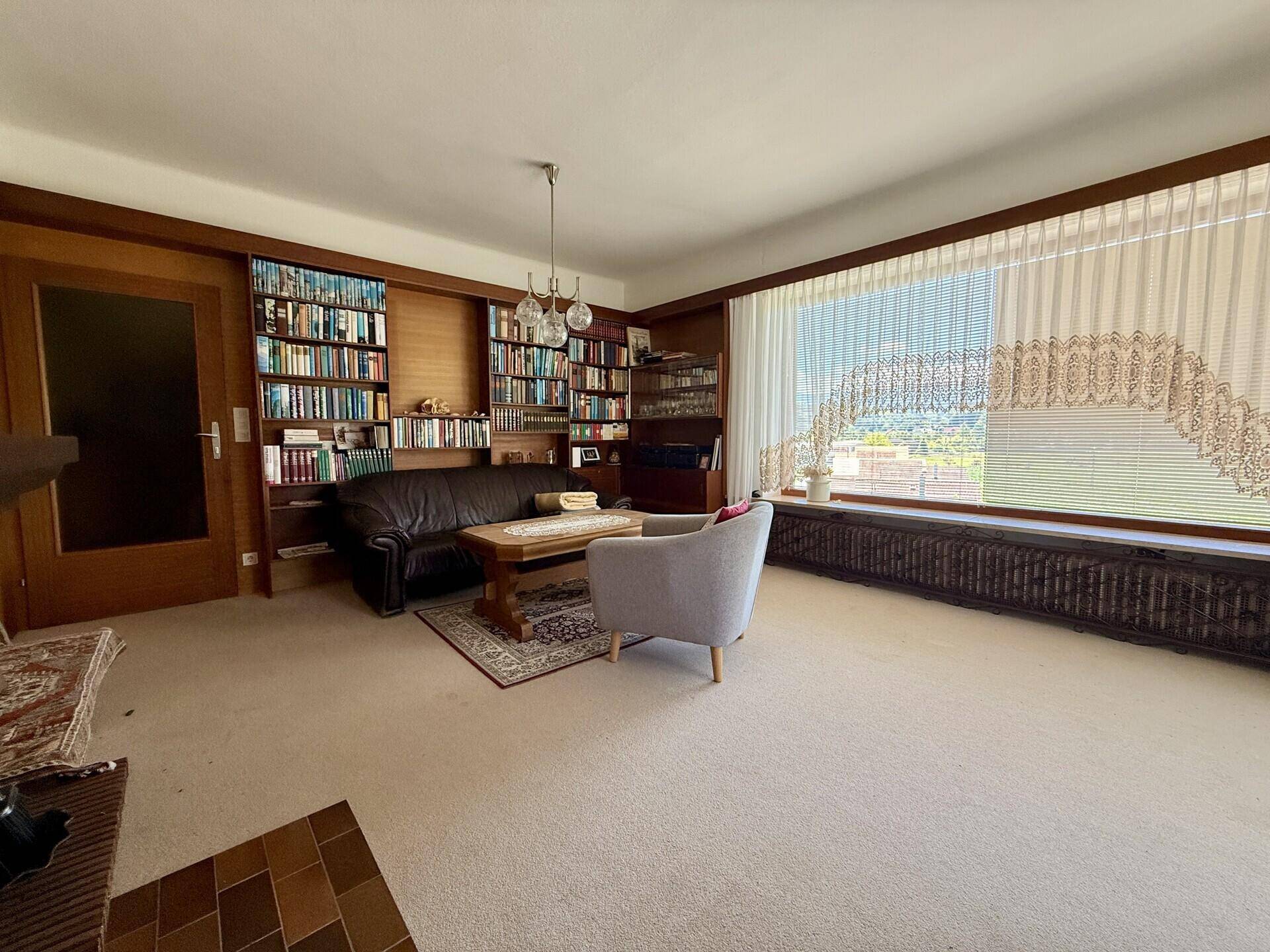 Gemütliches Wohnzimmer mit Teppichboden, Bücherregalen und einem großen Fenster mit Blick ins Grüne.