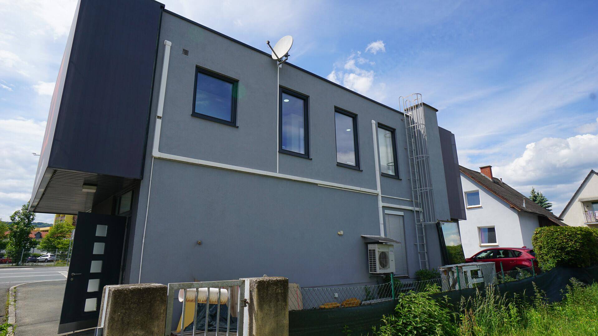 Seitenansicht des modernen Gebäudes mit grauer Fassade und mehreren Fenstern unter blauem Himmel.