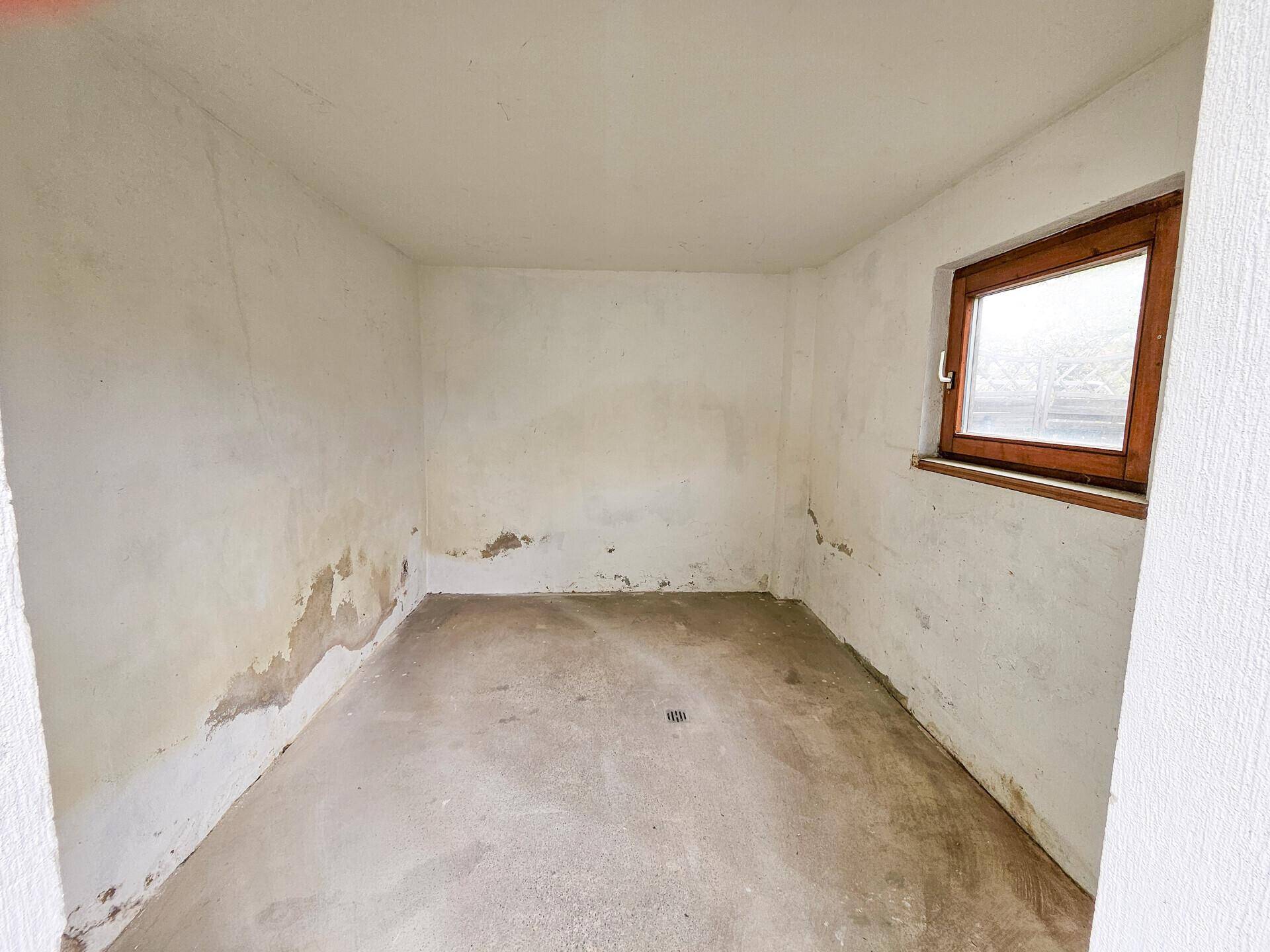 Leerer Abstellraum im Keller mit Fenster und sichtbaren Feuchtigkeitsspuren an den Wänden.