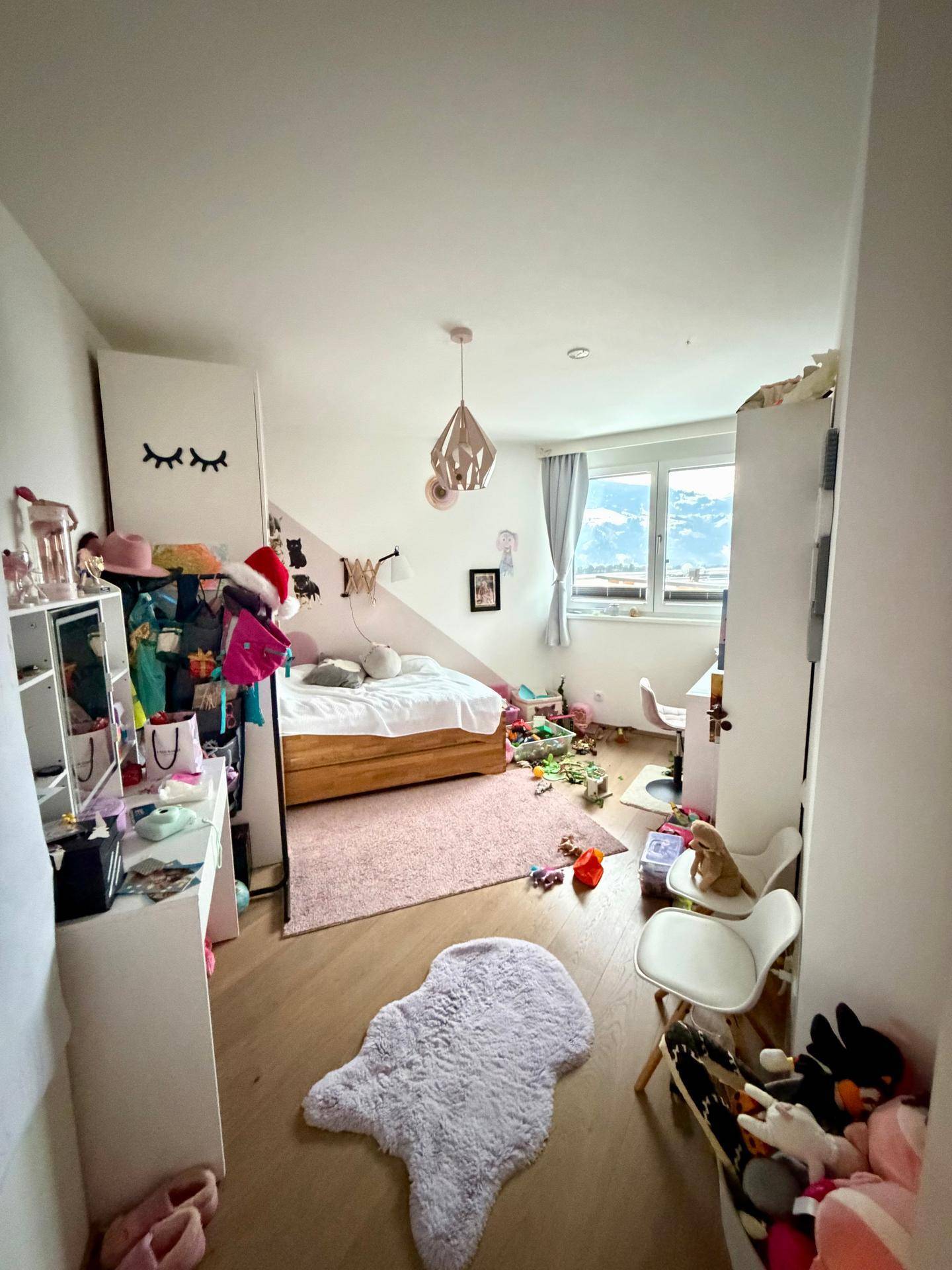 Mädchenzimmer mit Bett, Schreibtisch und Spielzeug, dekoriert in Rosa und Weiß mit Bergblick.
