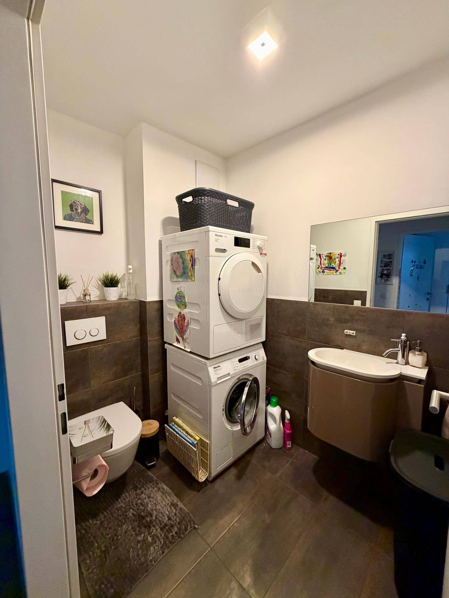 Kleines Badezimmer mit Toilette, Waschbecken und gestapelten Waschmaschinen, praktisch eingerichtet.