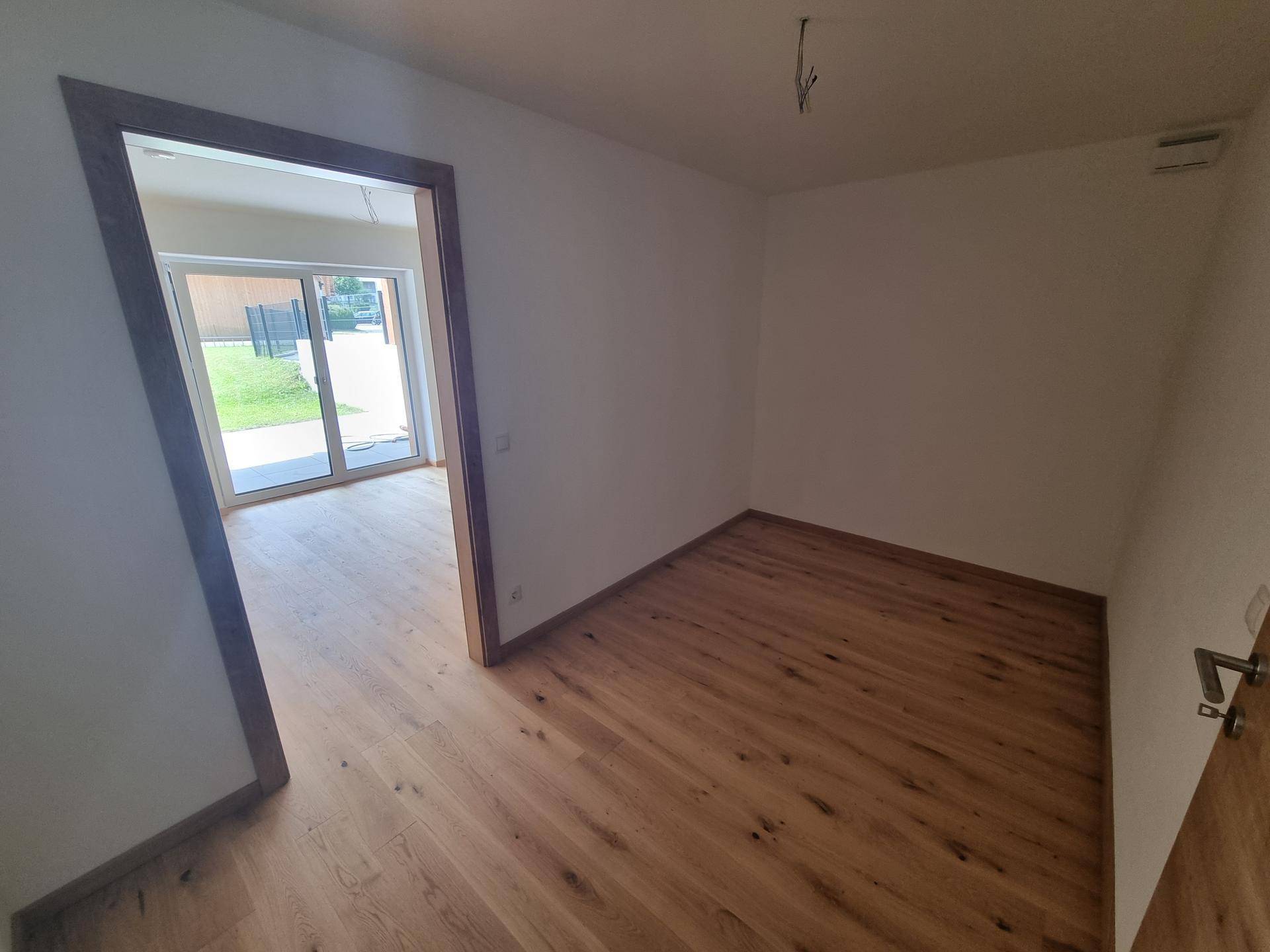 Heller, unmöblierter Raum mit Holzboden und Zugang zu einem weiteren Zimmer oder Außenbereich.