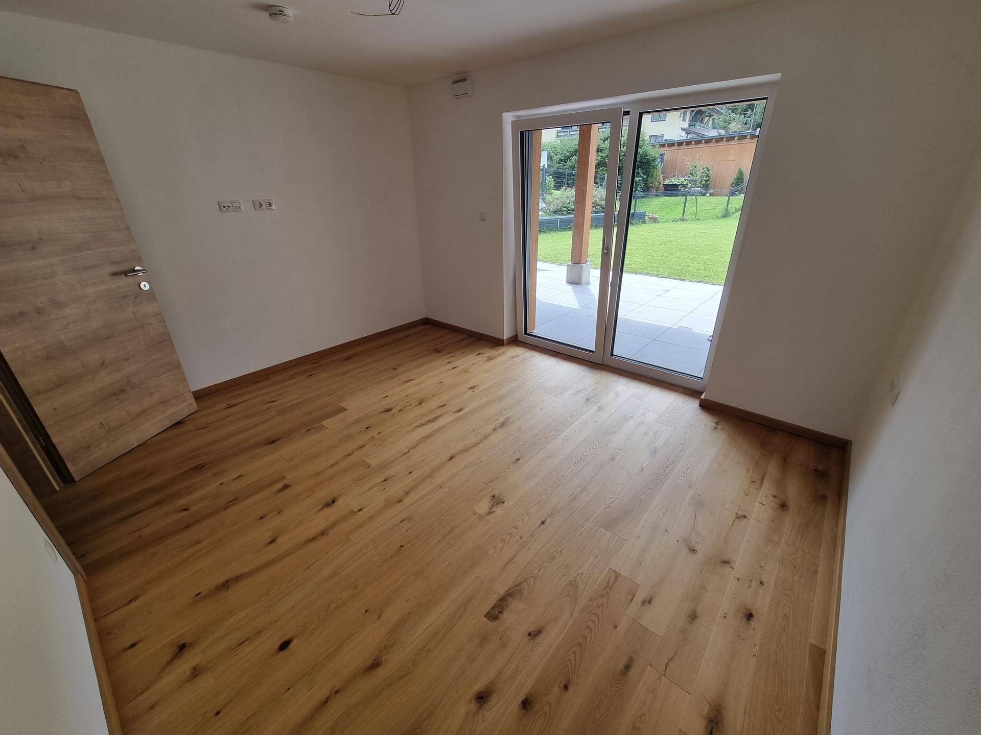 Geräumiger Raum mit Holzboden und großer Schiebetür, die zum Gartenbereich führt.