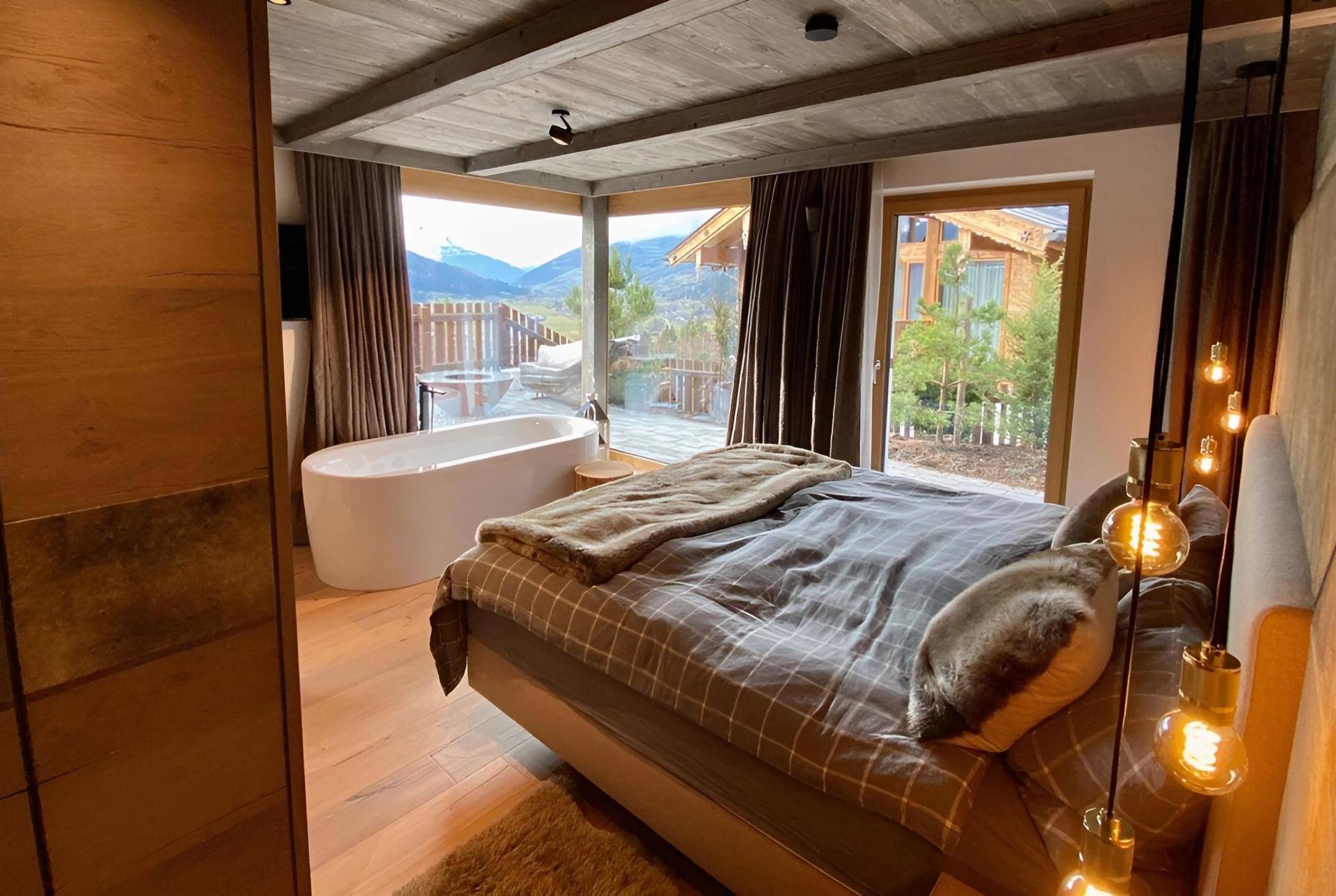 Helles Schlafzimmer mit Holzboden, freistehender Badewanne und Panoramablick auf die Berge.