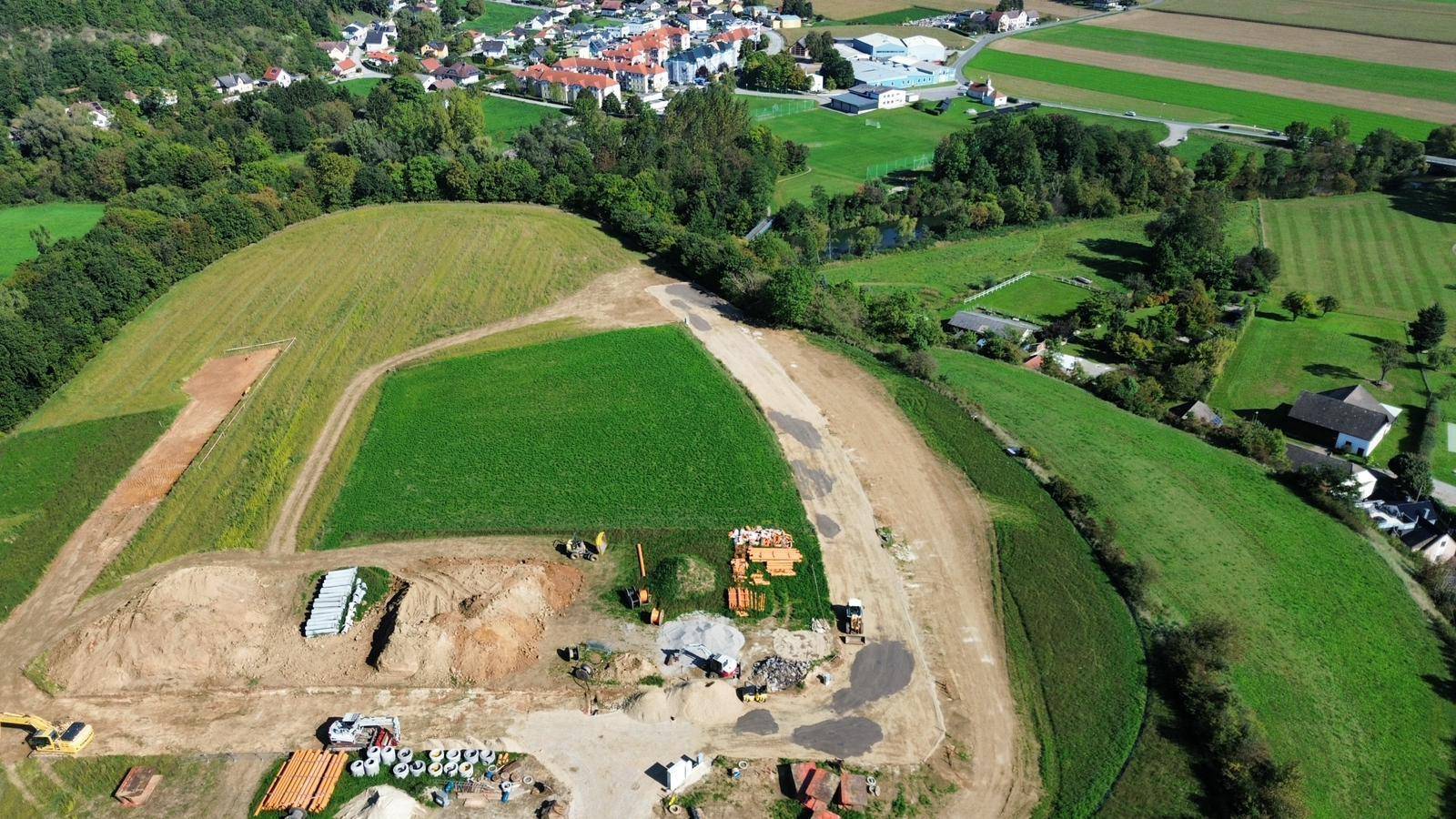 Luftaufnahme eines Baugebiets mit Erdhügeln, Baumaterialien und Maschinen in ländlicher Umgebung.